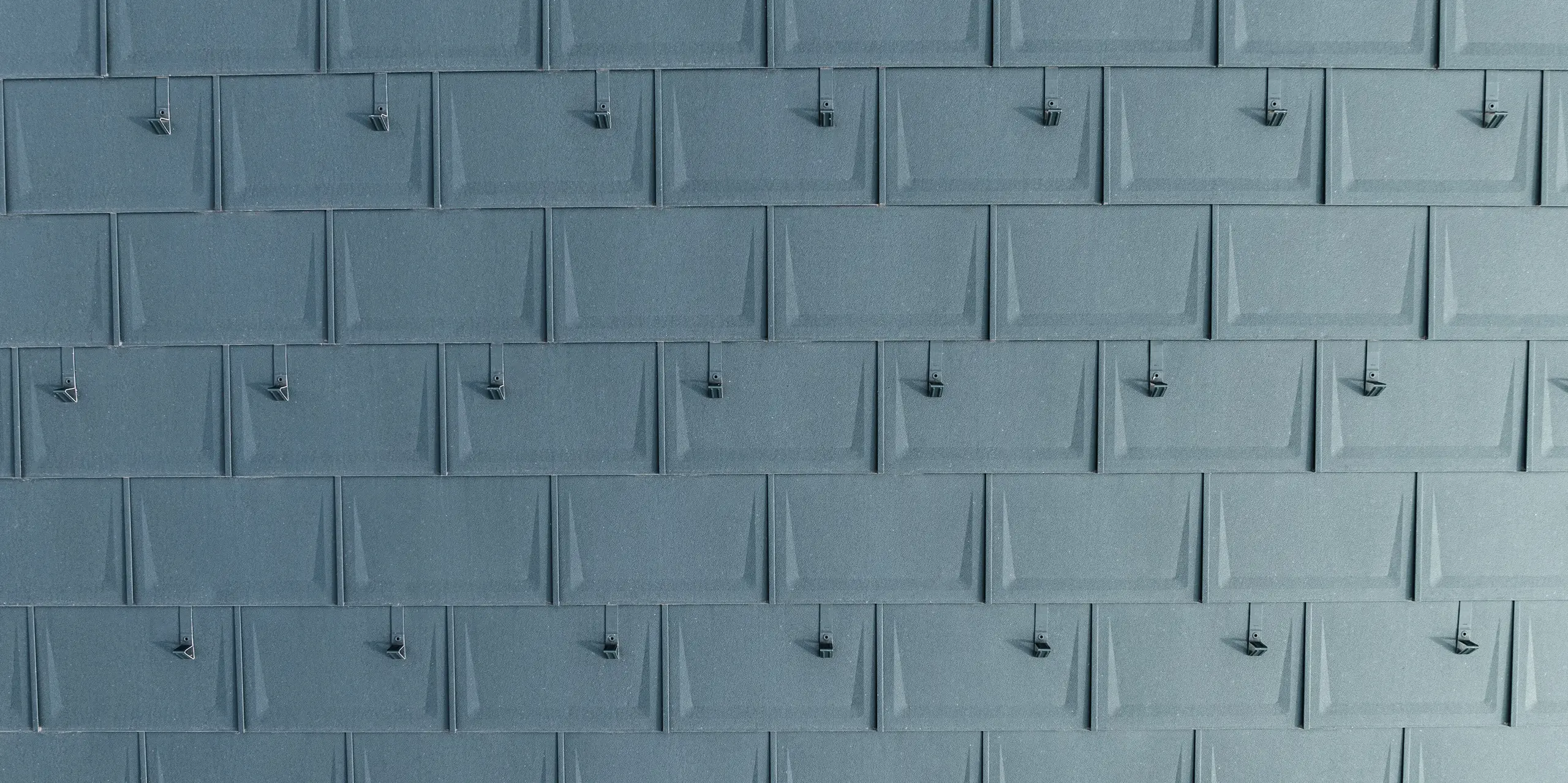 Detailný pohľad na presne položené strešné panely PREFA R.16 v antracitovej farbe P.10 s namontovanými snehovými zábranami. Lineárny hliníkový strešný panel zaujme svojím moderným vzhľadom, trvanlivosťou a extrémnou odolnosťou voči poveternostným vplyvom. Vďaka dômyselnej upevňovacej technológii je mimoriadne odolný proti búrke a rozbitiu. Snehové zábrany poskytujú dodatočnú bezpečnosť v zasnežených oblastiach a zabraňujú nekontrolovanému posúvaniu snehových más.