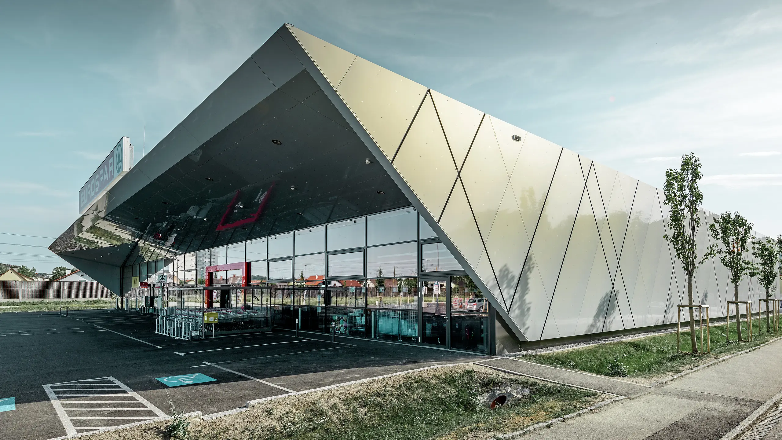 Vstupná časť supermarketu EUROSPAR kombinuje veľkorysé sklenené plochy s výraznou hliníkovou fasádou PREFA. Lesklý povrch v špeciálnej farbe Silver Gold podčiarkuje vysokú kvalitu a modernú architektúru tejto pobočky supermarketu. Hliníkové kompozitné panely sa v slnku lesknú v rôznych farbách v závislosti od uhla pohľadu.