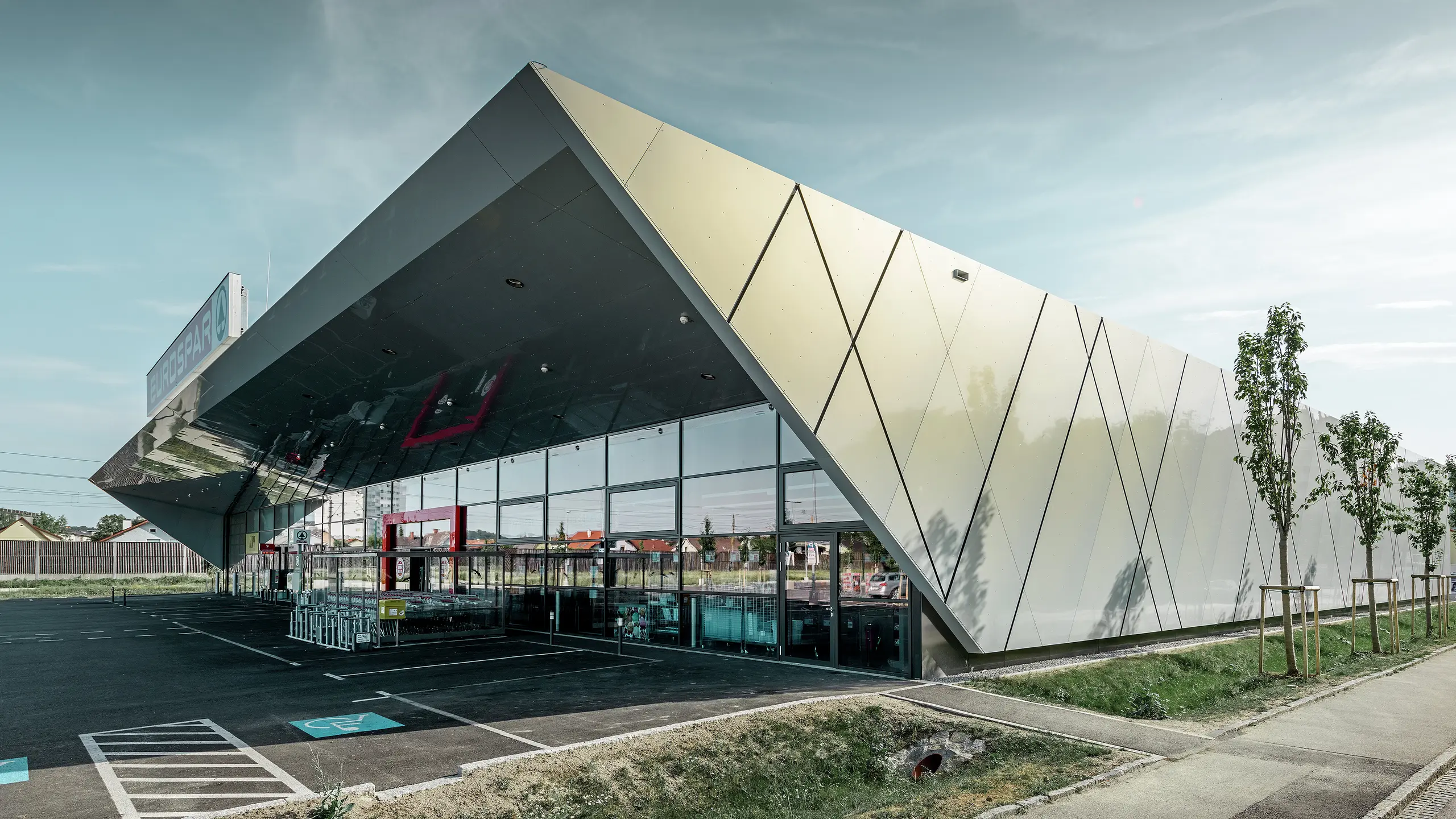 Vstupná časť supermarketu EUROSPAR kombinuje veľkorysé sklenené plochy s výraznou hliníkovou fasádou PREFA. Lesklý povrch v špeciálnej farbe Silver Gold podčiarkuje vysokú kvalitu a modernú architektúru tejto pobočky supermarketu. Hliníkové kompozitné panely sa v slnku lesknú v rôznych farbách v závislosti od uhla pohľadu.