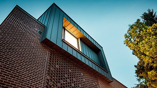 Záber zachytáva moderné riešenie fasády budapeštianskej viacgeneračnej vily z dynamickej žabie perspektívy. Vystupujúci kubus s hliníkom PREFALZ v odtieni P.10 antracit vytvára výrazný kontrast k tmavočerveným tehlám pod ním. Precízne spracovanie zvislých stojatých falcov zdôrazňuje čisté línie a vysokú kvalitu hliníkových produktov PREFA. Vďaka teplému svetlu vzniká silná hra materiálov, tieňov a kontrastov medzi kovom a tehlou, ktorá podčiarkuje inovatívny a nadčasový charakter budovy.