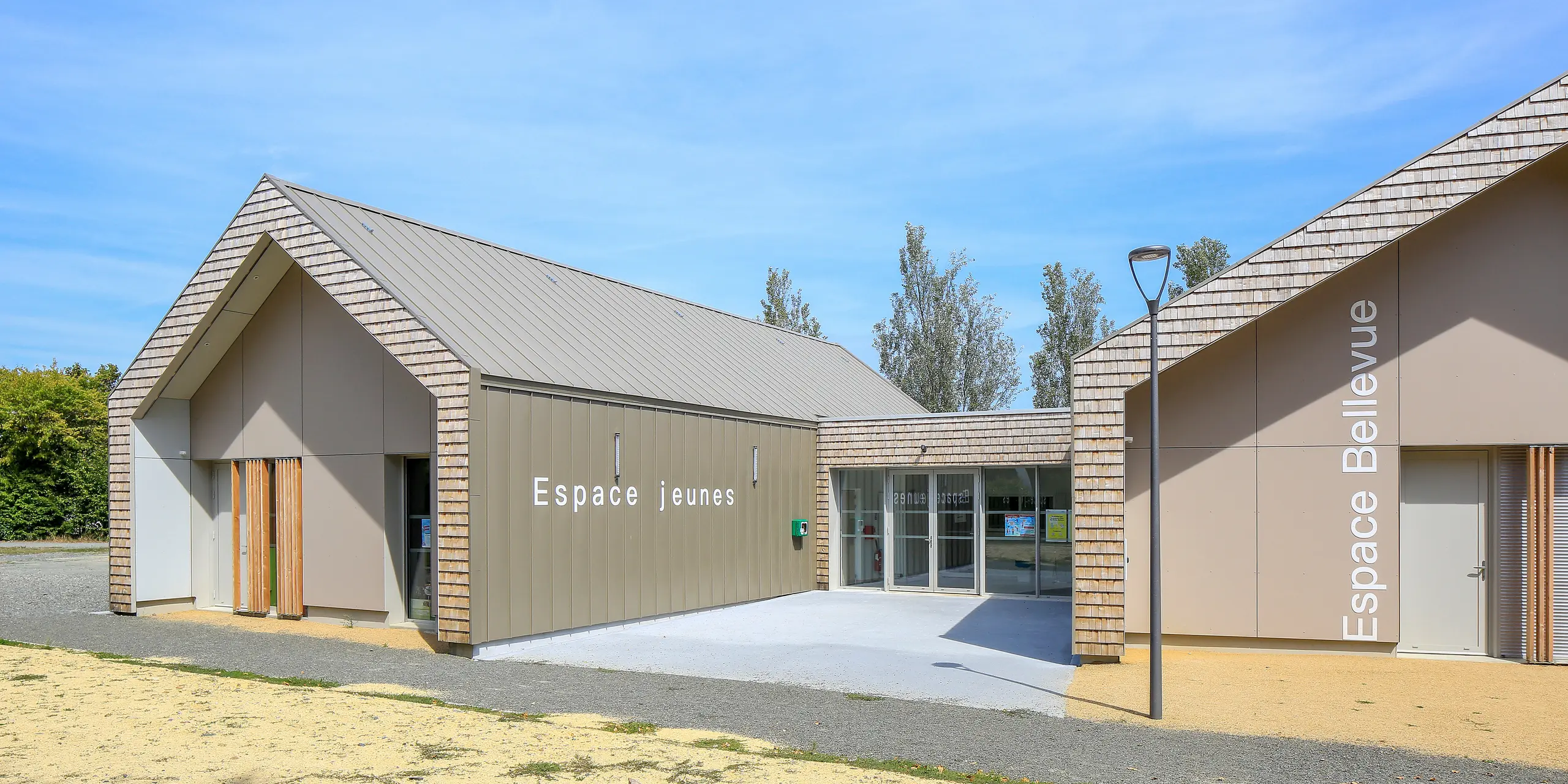 Salles Espace Bellevue v Mûrs-Erigné, Francúzsko, s nápismi „Espace Jeunes“ a „Espace Bellevue“ pred halou podujatia. Strešný a fasádny systém PREFALZ v bronzovej farbe zdobí veľkú časť moderného mládežníckeho centra. Hliníkové opláštenie na stojatú drážku od spoločnosti PREFA odolné voči poveternostným vplyvom ponúka trvanlivé a moderné riešenie pre stavebné projekty. Kombinácia hliníkového obkladu a prírodného vzhľadu dreva harmonicky zapadá do okolia.
