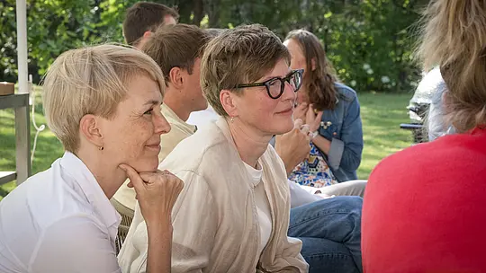 Detailný záber na Veroniku Roll (PREFA Marketing) a Julianu Croce (fotografka) počas diskusie odborníkov v rámci podujatia PREFARENZEN 2027 v záhrade vinárskej rezidencie. Obidve ženy sedia sústredene a pozorne a živou diskusiou rozoberajú moderné architektonické projekty. Scéna ponúka prirodzené osvetlenie, letnú zeleň a uvoľnenú atmosféru. V pozadí sú viditeľné hliníkové produkty PREFA, ktoré symbolizujú trvácne, esteticky príťažlivé strechy a fasády, v ktorých sa spája funkčnosť s dizajnom. Obrázok zdôrazňuje kreatívnu výmenu názorov a intenzívnu diskusiu poroty o dizajnových nápadoch, ako aj inšpiráciu, ktorú prinášajú vysoko kvalitné, inovatívne hliníkové riešenia PREFA pre architektonické a stavebné projekty.