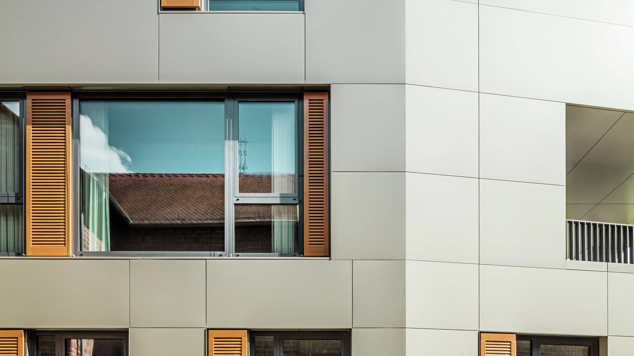 Detailný záber fasády radnice v Alzenau v Nemecku s hliníkovými kompozitnými doskami PREFA v bronzovej farbe. Veľké okná sú orámované elegantnými drevenými žalúziami v teplej hnedej farbe, ktoré dodávajú fasáde príjemnú textúru. V popredí je vidieť upravený kvetinový záhon, ktorý dopĺňa mestský dizajn budovy zelenými akcentmi. Jasná obloha a čisté línie architektúry podčiarkujú moderný charakter budovy.