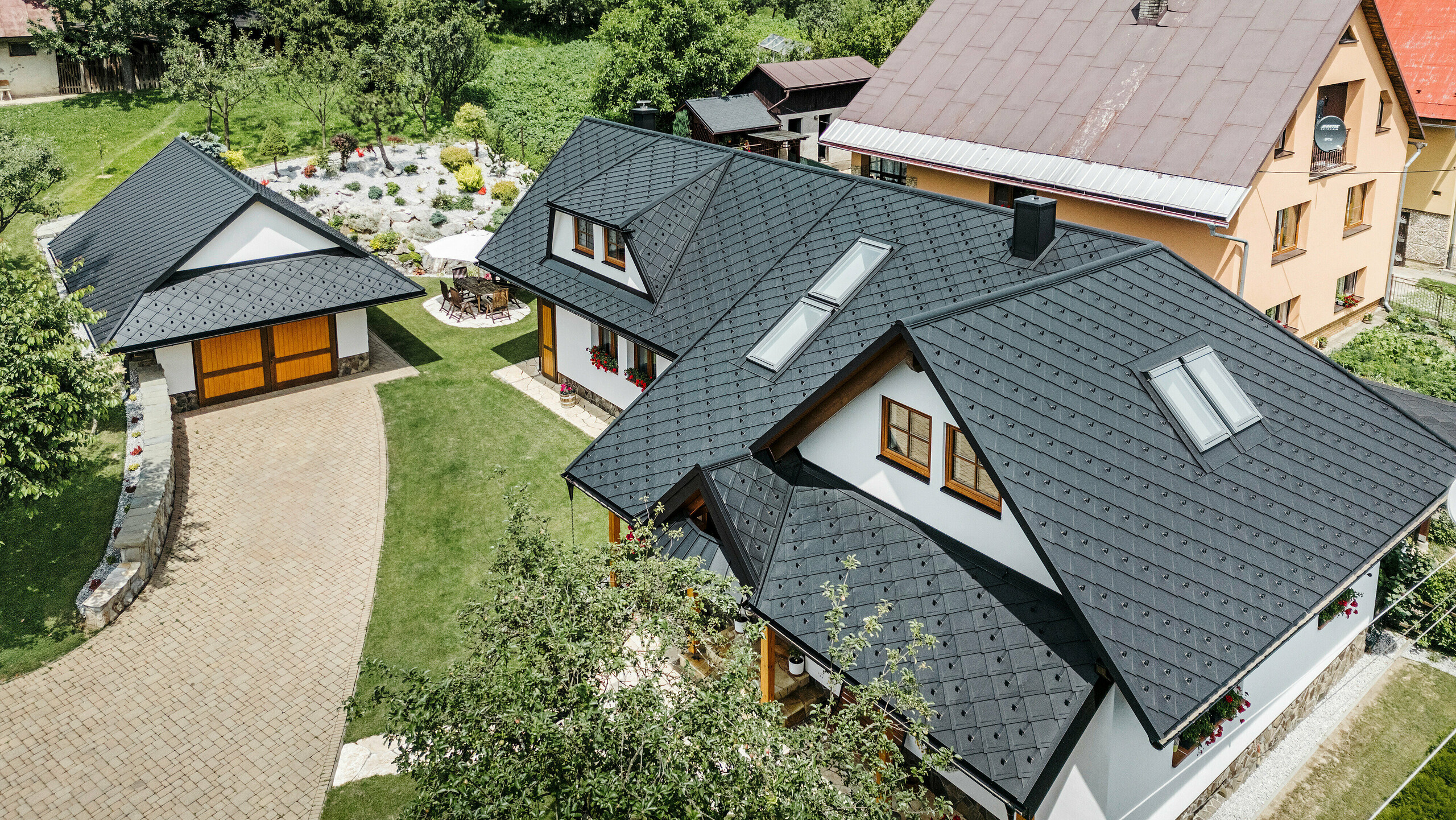 Letecký pohľad na tradičný rodinný dom v Starej Bystrici na Slovensku, pokrytý strešnými šablónami PREFA 29 × 29 vo farbe P.10 čierna. Strecha zaujme elegantným pultovým vzhľadom a harmonickým začlenením vikierov a strešných okien. Okolitá zeleň, udržiavaná záhrada a vydláždená príjazdová cesta podčiarkujú príťažlivý celkový vzhľad budovy.