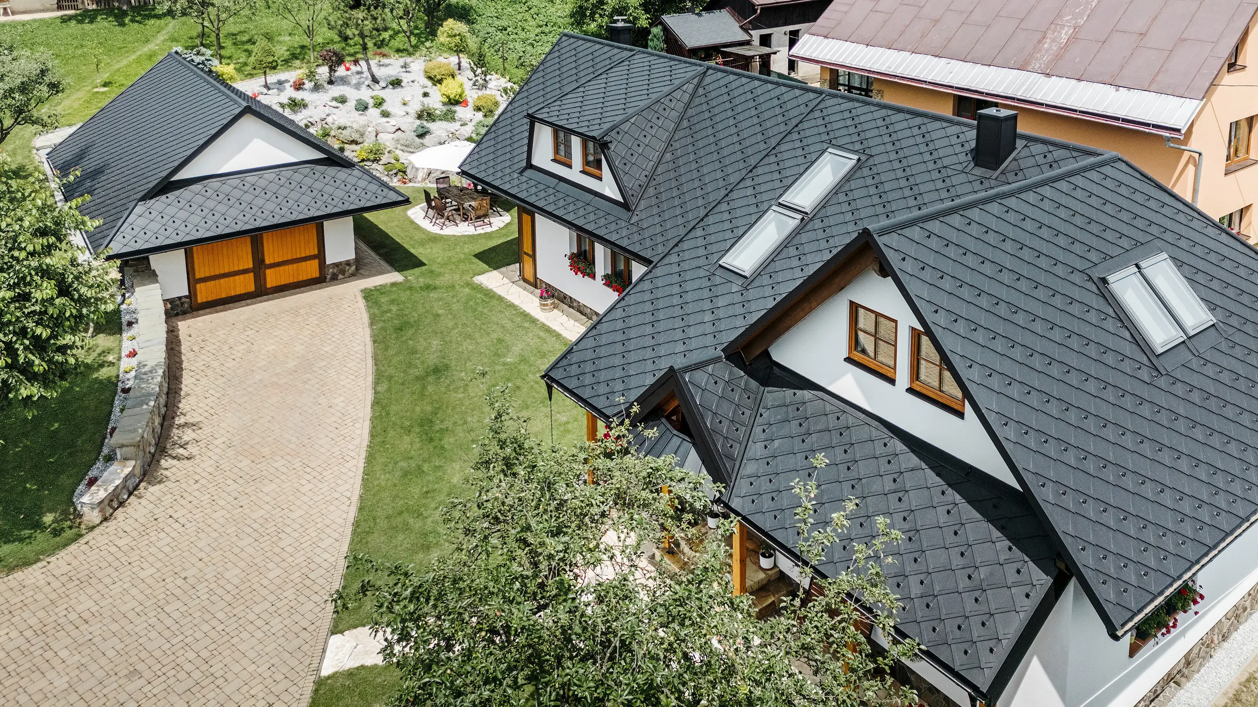 Letecký pohľad na tradičný rodinný dom v Starej Bystrici na Slovensku, pokrytý strešnými šablónami PREFA 29 × 29 vo farbe P.10 čierna. Strecha zaujme elegantným pultovým vzhľadom a harmonickým začlenením vikierov a strešných okien. Okolitá zeleň, udržiavaná záhrada a vydláždená príjazdová cesta podčiarkujú príťažlivý celkový vzhľad budovy.