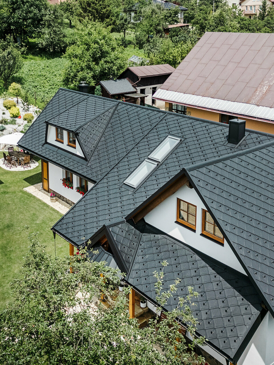 Letecký pohľad na tradičný rodinný dom v Starej Bystrici na Slovensku, pokrytý strešnými šablónami PREFA 29 × 29 vo farbe P.10 čierna. Strecha zaujme elegantným pultovým vzhľadom a harmonickým začlenením vikierov a strešných okien. Okolitá zeleň, udržiavaná záhrada a vydláždená príjazdová cesta podčiarkujú príťažlivý celkový vzhľad budovy.