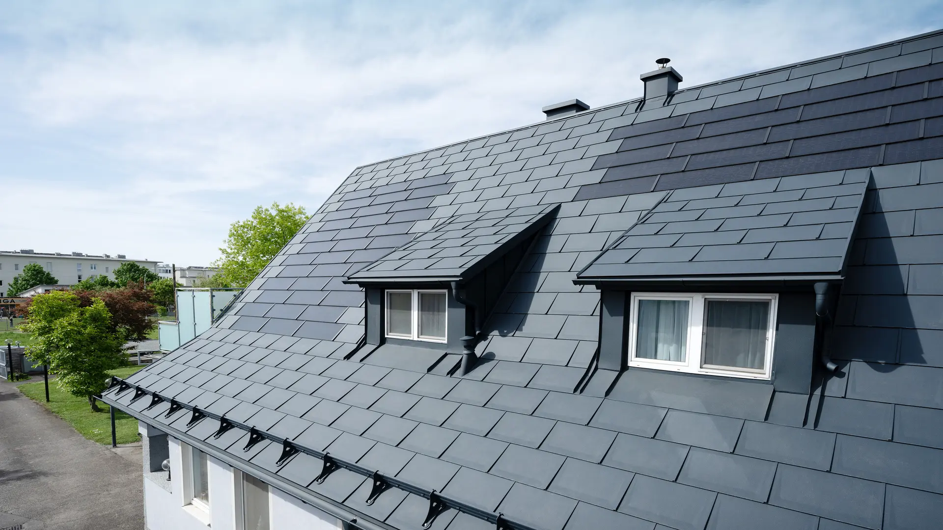 Detailný záber strechy pokrytej veľkými solárnymi strešnými panelmi PREFA a strešnými panelmi PREFA R.16 v odtieni P.10 antracitová. Viditeľné sú dve vikiery. Solárne strešné panely sú rovnomerne rozmiestnené po streche a vytvárajú moderné a udržateľné riešenie.