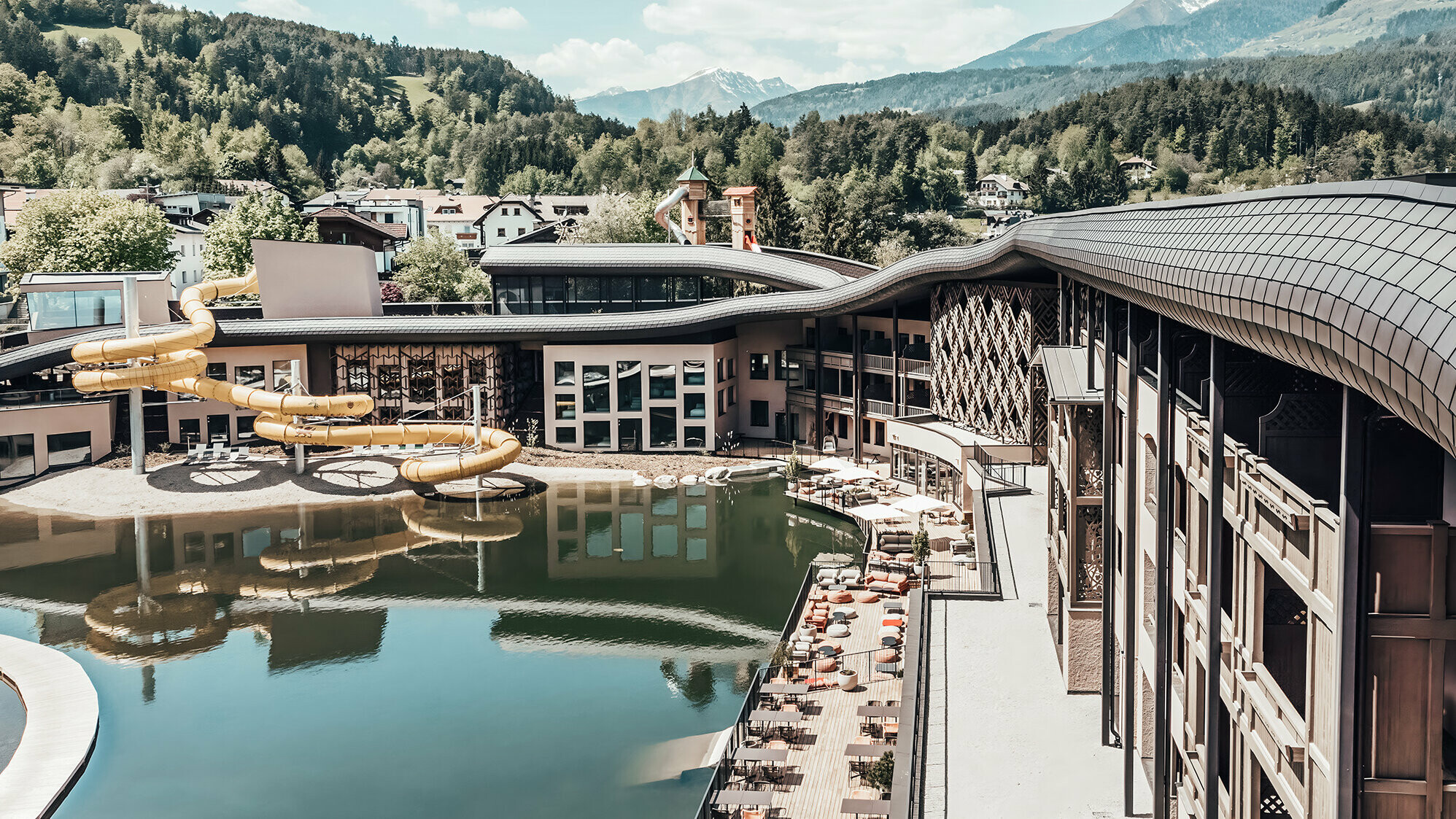 Überblick auf das Hotel mit See und Wasserrutsche. Im Hintergrund ist hügelige und bewaldete Fläche zu sehen.