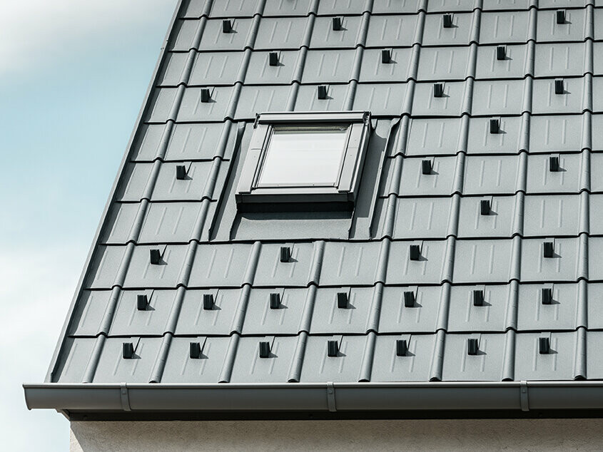 Falcovaná škridla PREFA s protisnehovými zachytávačmi v antracitovej farbe, položená so zabudovaným strešným oknom a dažďovým žľabom PREFA. Strešná plocha s plechovou strechou PREFA