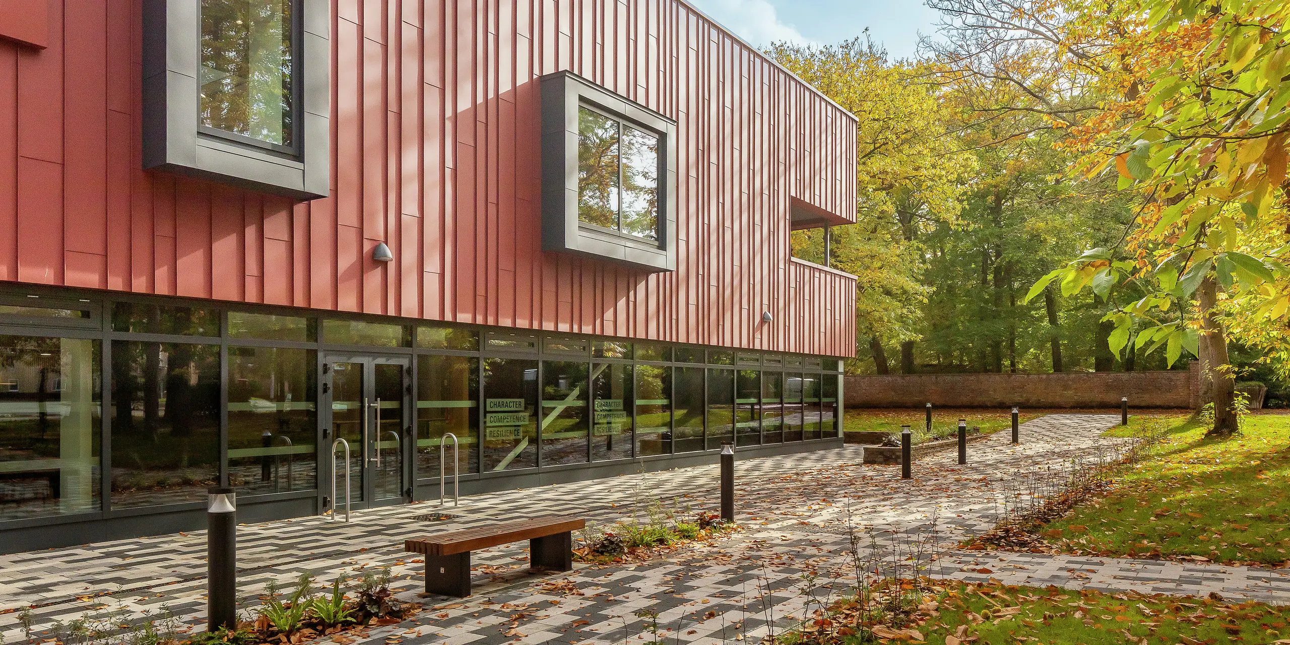 Harmonická architektúra v zeleni: Moderná stojatá falcová fasáda Soldiers' Centre v Pirbright je vyrobená z PREFALZu v odtieni P.10 tmavočervená. Robustný hliníkový obklad sa perfektne hodí do jesenného prostredia. Nápadné, vyčnívajúce okenné rámy v P.10 Anthrazit dodávajú štýlové akcenty. Veľké sklenené plochy na prízemí zaisťujú transparentnosť a otvorenosť. Dláždené predsieň s lavičkou láka k oddychu – úspešné spojenie dizajnu, funkčnosti a prírody.