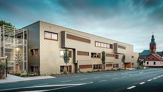 Záber z úrovne ulice zobrazuje novú materskú školu v Alzenau v pôsobivom dialógu s historickým mestským prostredím, vrátane výrazného kostola v pozadí. Moderná fasáda z bronzovo sfarbených PREFA fasádnych šablón 44 × 44 dodáva budove elegantný, takmer sochársky výraz. Precízne umiestnené okná, vertikálne drevené lamely a posunuté plochy fasády vytvárajú hĺbku a rytmus. Na ľavej strane je viditeľné filigránske schodisko s bielou kovovou konštrukciou, ktoré pôsobí moderným a ľahkým dojmom. Jasný tvaroslovný jazyk, udržateľný výber materiálov a kvalitná hliníková fasáda robia z budovy vzorový projekt urbanistickej integrácie a perspektívnej výstavby s fasádnymi systémami PREFA.