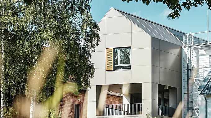 Na obrázku je moderná radnica v Alzenau, ktorej fasáda je vyrobená z hliníkových kompozitných dosiek PREFA v elegantnom bronzovom odtieni. Špicatá strecha a veľké okná zdôrazňujú moderný charakter budovy. V popredí budovu obklopujú stromy a zeleň, pričom mäkké osvetlenie a jasná obloha podčiarkujú moderný mestský priestor.