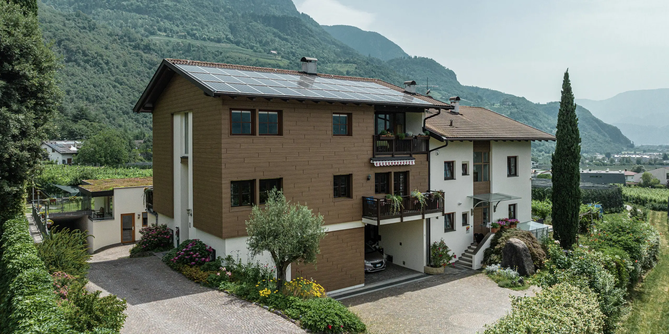Renovované rekreačné apartmány Prechtlhof v Merane, Južné Tirolsko, s obkladmi PREFA v orechovo hnedom vyhotovení a fotovoltickou inštaláciou na streche. Odolná hliníková fasáda v drevenom vzhľade zaisťuje prirodzenú estetiku, najvyššiu odolnosť a nízke náklady na údržbu, čo je ideálne pre alpské prostredie.