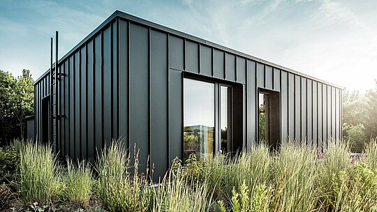 Záber zachytáva modernú, kubickú strešnú terasu budapeštianskej viacgeneračnej vily, obloženú kvalitným hliníkom PREFALZ v odtieni P.10 antracit. Jasne štruktúrované stojaté falce vytvárajú elegantné vertikálne línie, ktoré jemné slnečné svetlo ešte viac zvýrazňuje. Obklopená hustou prírodnou zeleňou vzniká zaujímavý kontrast medzi modernou kovovou architektúrou a bujnou vegetáciou. Trvácne a poveternostným vplyvom odolné hliníkové produkty PREFA podčiarkujú vysokú kvalitu vyhotovenia a robia zo strešnej terasy výrazný architektonický prvok.