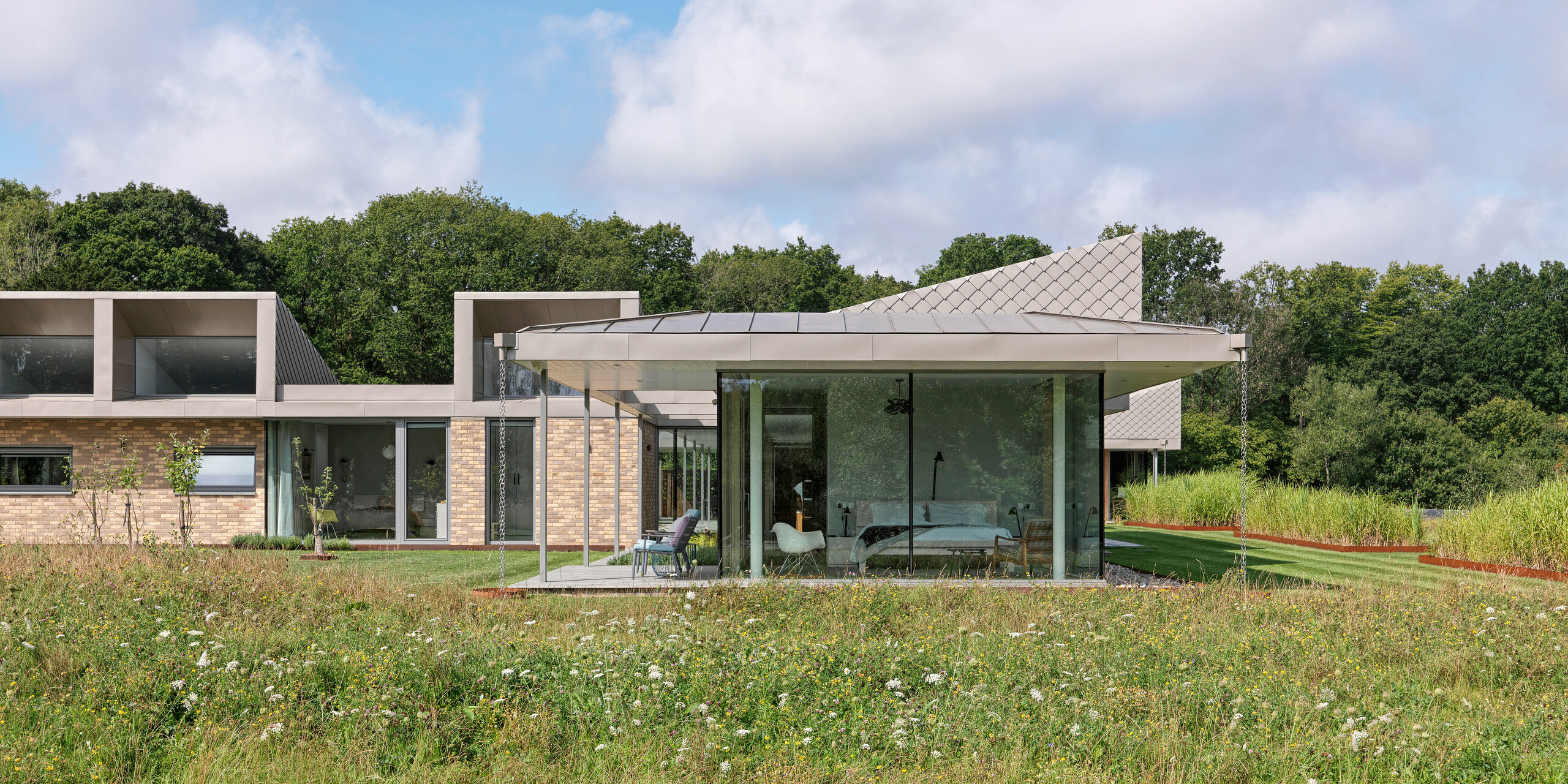 Bočný pohľad na moderný bungalov v West Sussex s hliníkovou strechou PREFALZ a akcentovanými fasádnymi plochami z PREFA šablón 20 × 20 v odtieni P.10 bronzová. Sklenené priečelie obývacích priestorov siahajúce až po strop sa otvára na lúku s divokými kvetmi v popredí a vytvára plynulé prepojenie medzi interiérom a prírodou. Odolný strešný a fasádny systém PREFA poskytuje budove nielen ochranu, ale aj architektonickú eleganciu.