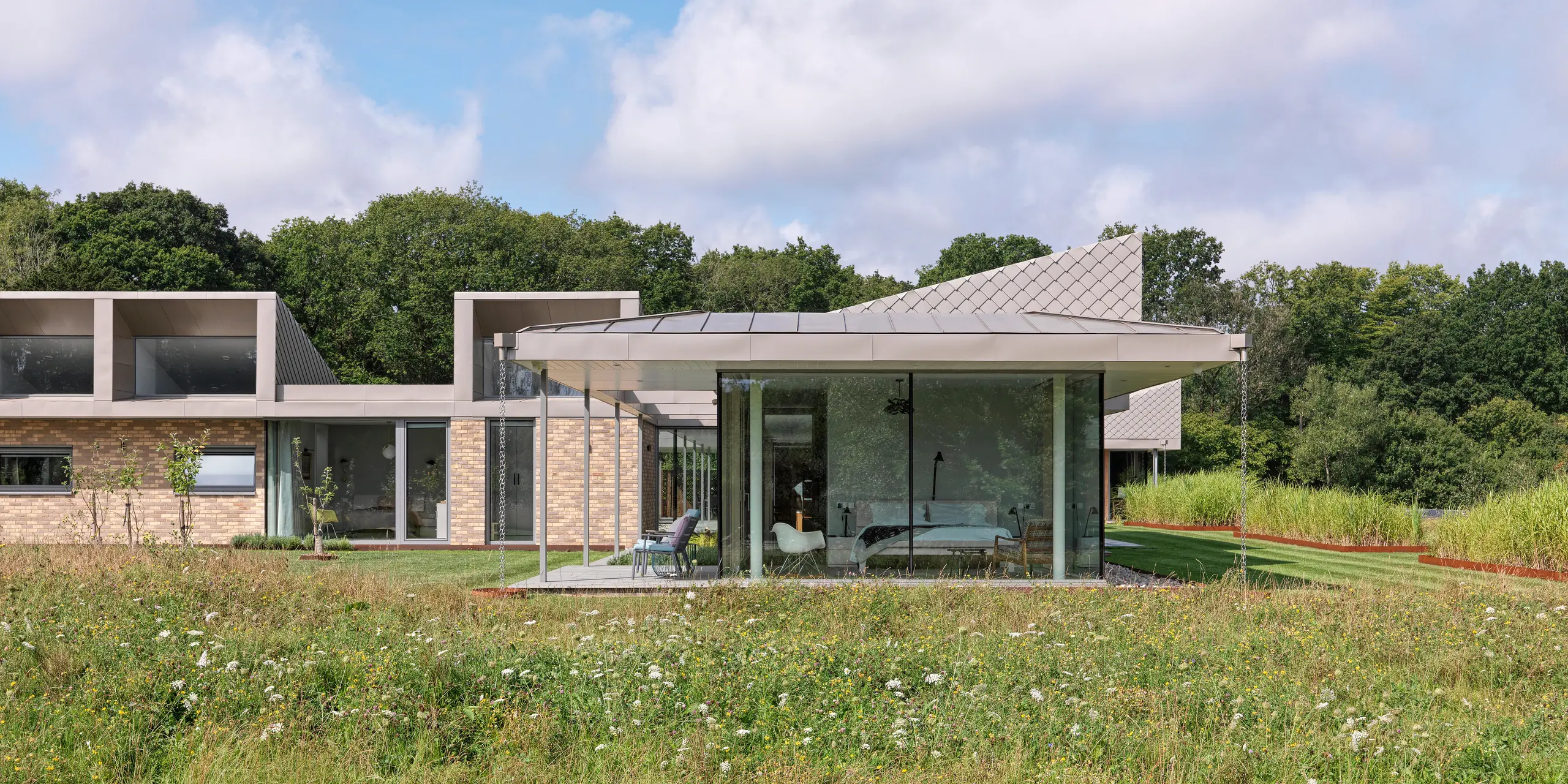 Bočný pohľad na moderný bungalov v West Sussex s hliníkovou strechou PREFALZ a akcentovanými fasádnymi plochami z PREFA šablón 20 × 20 v odtieni P.10 bronzová. Sklenené priečelie obývacích priestorov siahajúce až po strop sa otvára na lúku s divokými kvetmi v popredí a vytvára plynulé prepojenie medzi interiérom a prírodou. Odolný strešný a fasádny systém PREFA poskytuje budove nielen ochranu, ale aj architektonickú eleganciu.
