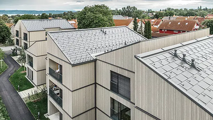 Letecký pohľad na moderné bytové domy v Utting am Ammersee, Nemecko. Obytný projekt zaujme inovatívnou hybridnou konštrukciou z dreva a odolnými strešnými panelmi PREFA R.16 v tmavosivej farbe P.10. Budovy harmonicky zapadajú do zeleného okolia a ponúkajú estetickú kombináciu drevených fasád a vysokokvalitnej hliníkovej konštrukcie strechy. Precízna montáž strešných panelov zabezpečuje odolnosť voči poveternostným vplyvom a trvanlivosť, zatiaľ čo čisté línie a moderná architektúra vytvárajú prívetivé a udržateľné obytné prostredie.