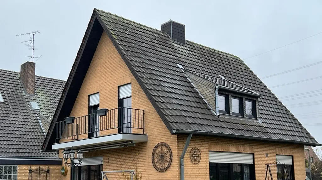 Rodinný dom zo žltého klinkeru so strmou sedlovou strechou a tmavou strešnou krytinou pred renováciou strechy; na strane záhrady s balkónom, vikierom, terasou s posedením a trávnikom v popredí.