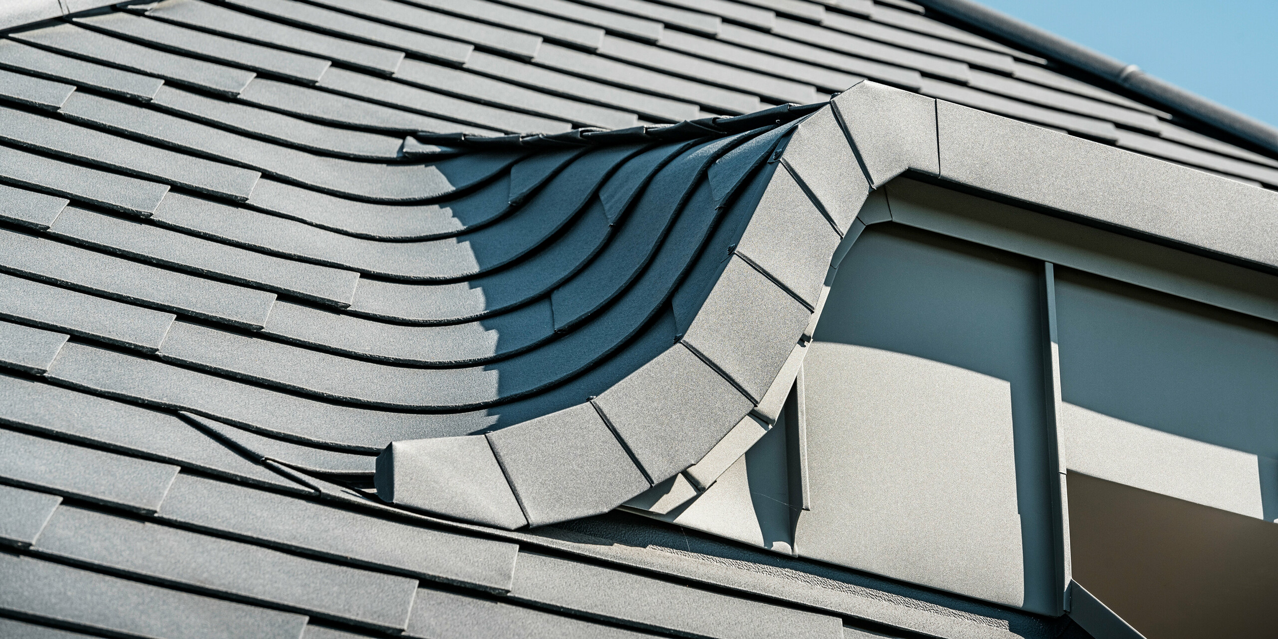 Detailný záber na strechu so zakrivenými vikiermi pokrytými strešnými šindľami PREFA v antracitovej farbe P.10