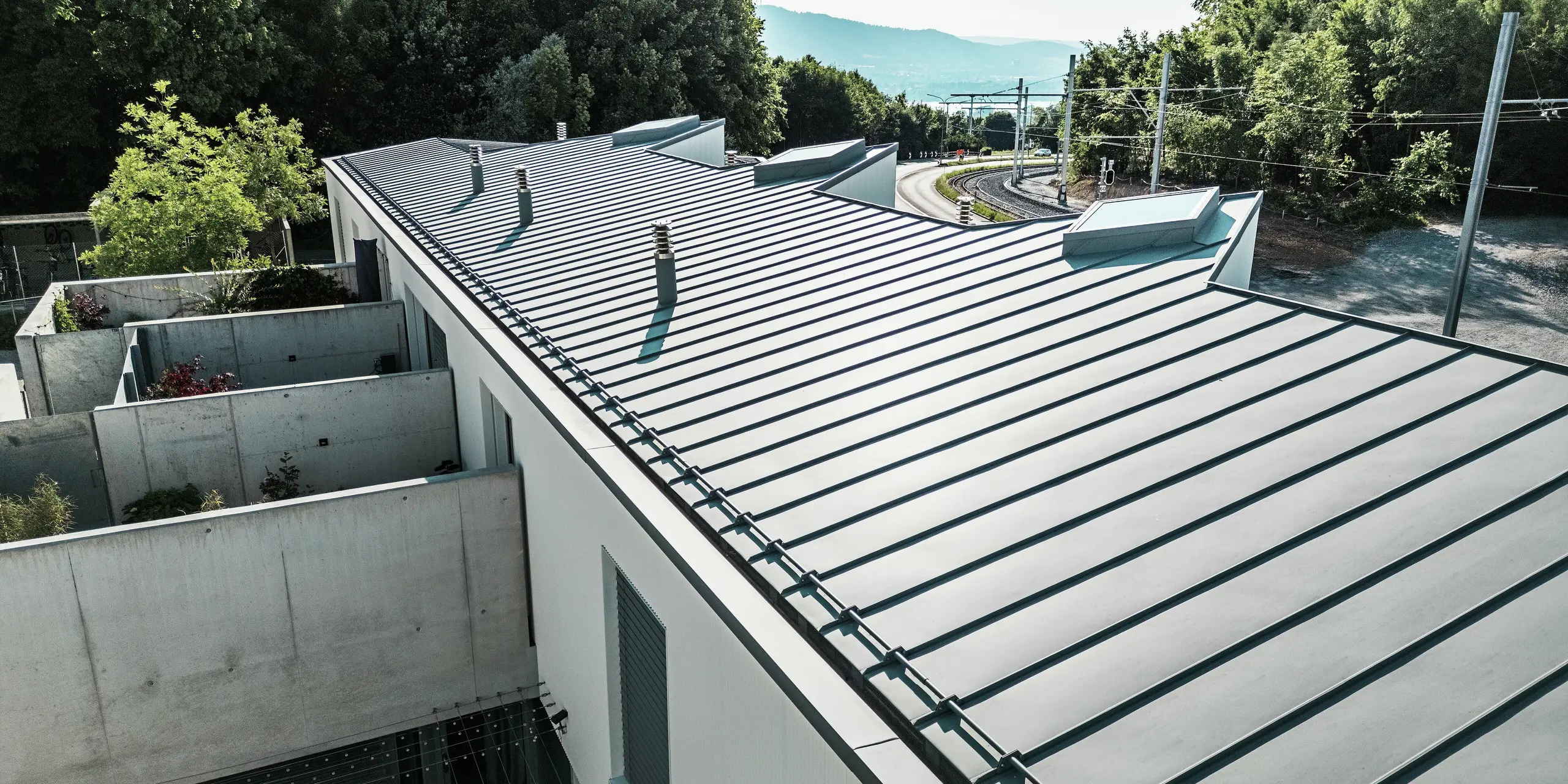 Langgezogene Dachansicht eines Tiny Homes Wohnkomplexes in Zollikerberg mit hochwertigem PREFALZ Stehfalzdach in Patinagrau. Das witterungsbeständige PREFA Aluminiumdach passt sich harmonisch der modernen Architektur und der naturnahen Umgebung an. Die klare Linienführung, integrierten Dachaufbauten und die nachhaltige Materialwahl machen dieses Dach zur langlebigen Premiumlösung.