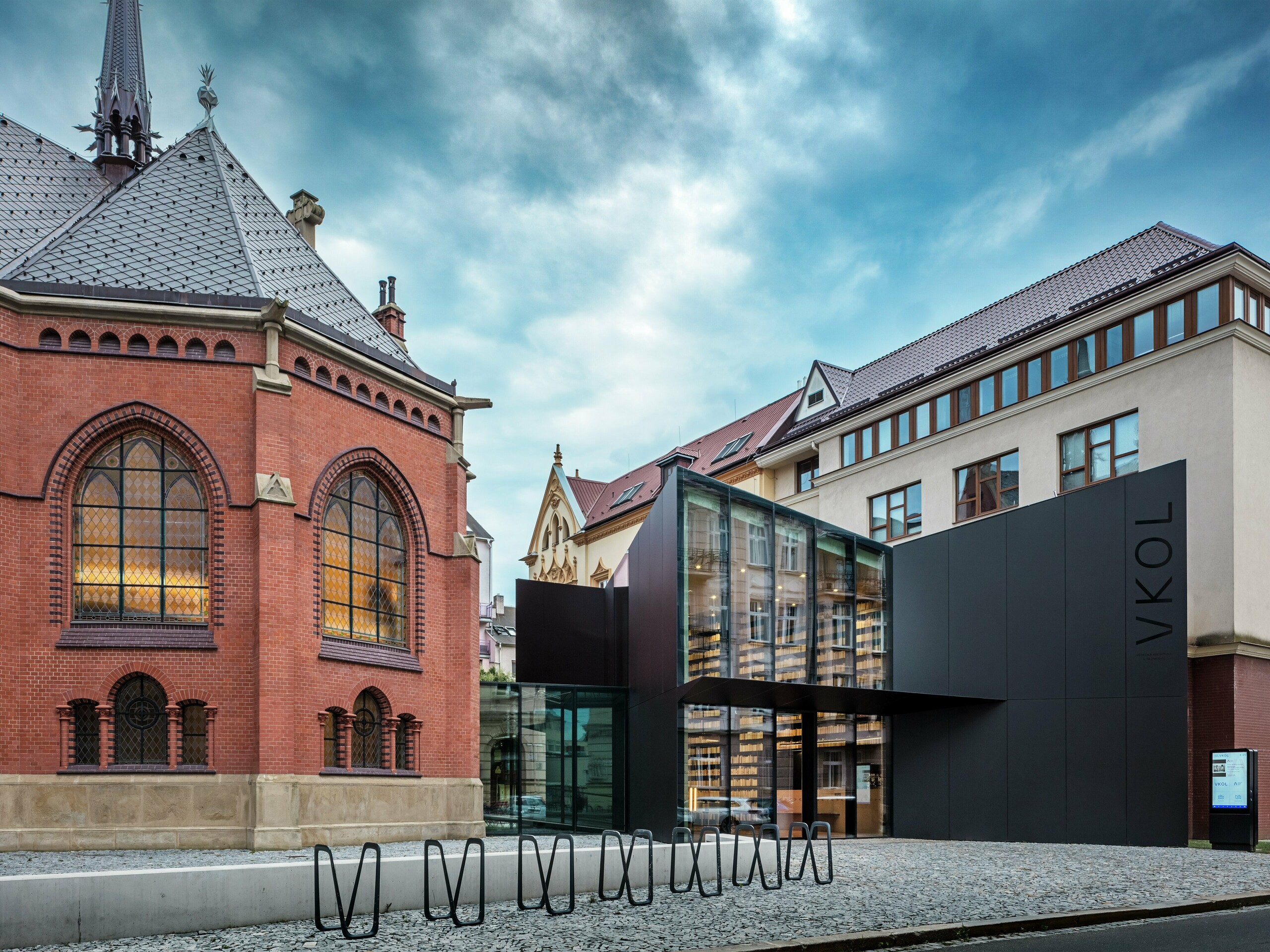 Obrázok ukazuje fascinujúce spojenie historickej a súčasnej architektúry v Olomouci. Na ľavej strane stojí majestátny Červený kostol s charakteristickými gotickými oknami a červenými tehlovými múrmi, zatiaľ čo nová budova na pravej strane kontrastuje s modernou čiernou fasádou z hliníkových kompozitných dosiek PREFABOND. Sklenená fasáda novej budovy poskytuje pohľad na inovatívny dizajn knižnice a dodáva komplexu elegantný, otvorený charakter. Presné spracovanie dosiek PREFABOND zaisťuje jednotný vzhľad, ktorý harmonicky zapadá do historického prostredia a zároveň vytvára moderný dojem. Architektúra tohto projektu dokonale spája tradíciu a inováciu, pričom materiály nielenže presviedčajú esteticky, ale aj zaisťujú dlhú životnosť a udržateľnosť.