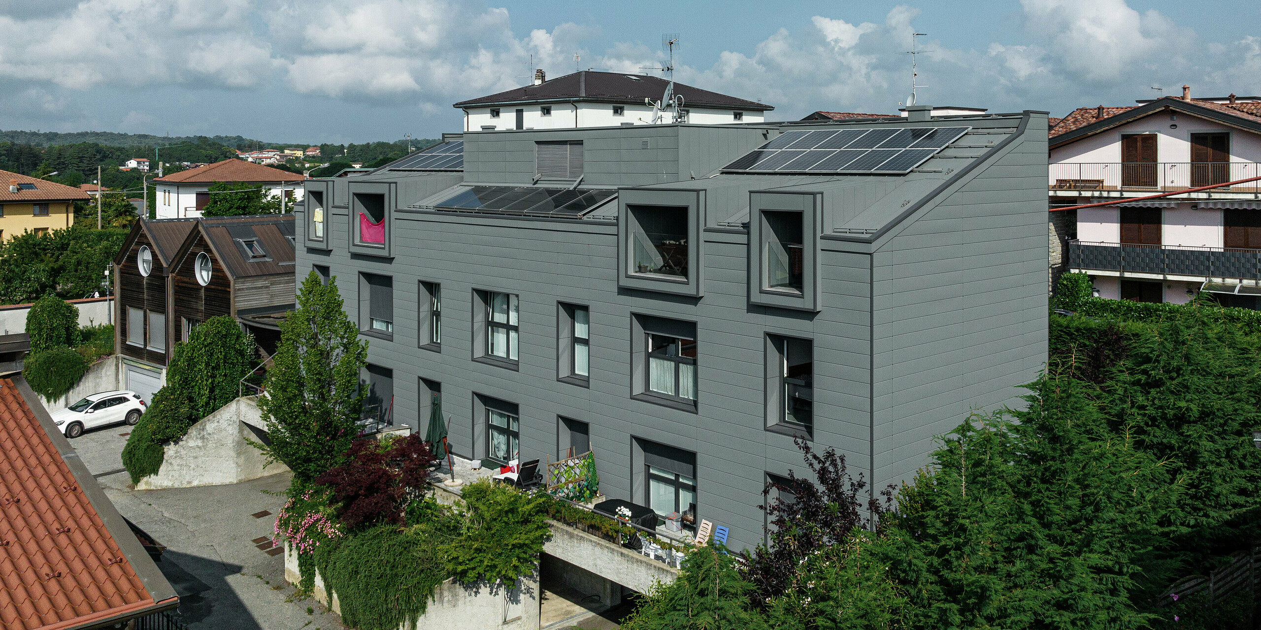 Priestranný pohľad na moderný obytný komplex v talianskom meste Faloppio s výraznou fasádou PREFA Sidings a energeticky účinnou strechou s fotovoltickými modulmi. Táto kombinácia inovatívnej architektúry a udržateľnej výroby energie je vynikajúcim príkladom modernej a udržateľnej výstavby. Plechová fasáda zabezpečuje odolnosť a minimalizuje údržbu, zatiaľ čo solárne články poskytujú ekologický zdroj elektrickej energie.