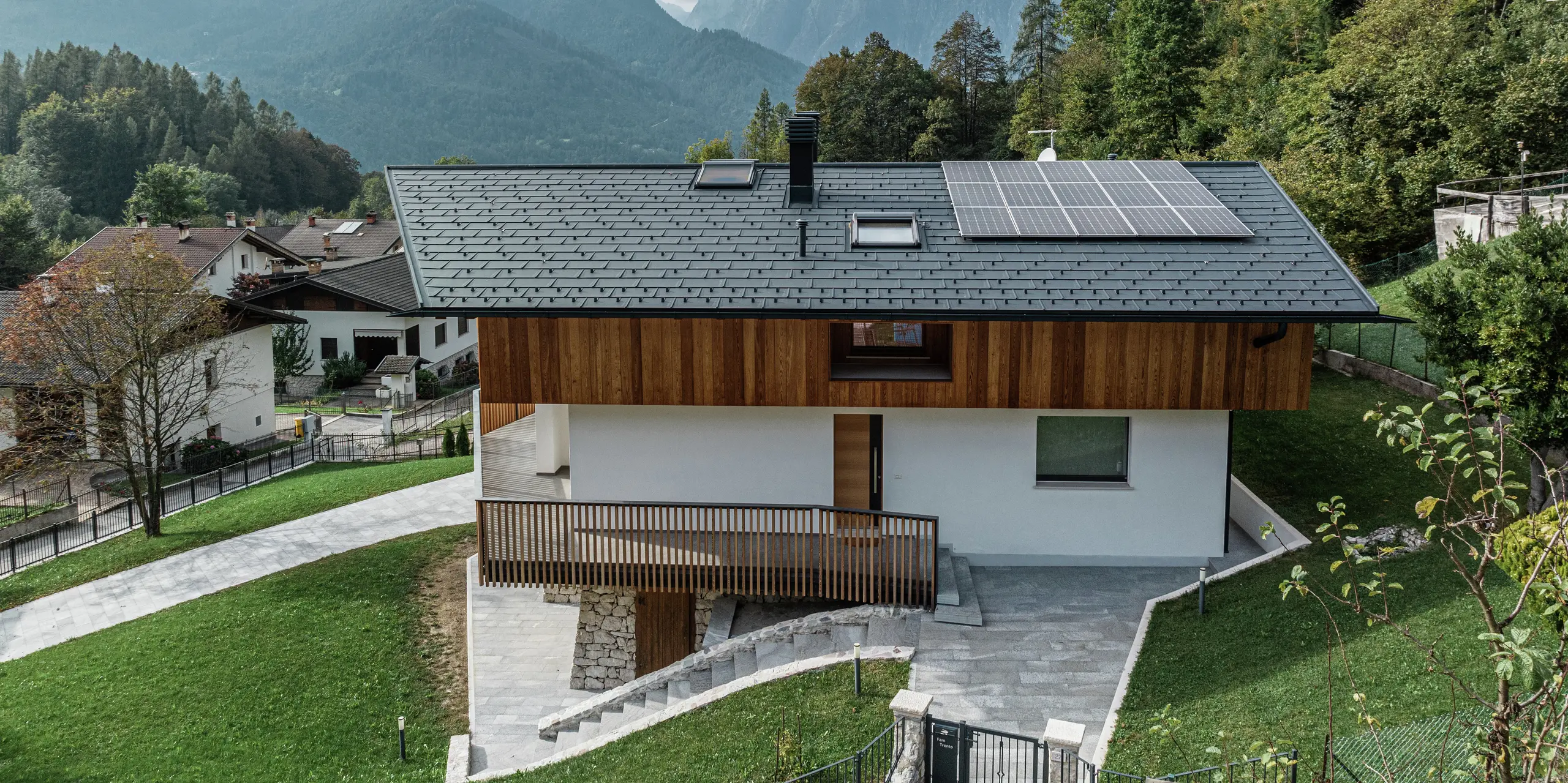 Bočný pohľad na moderný rodinný dom v Agorde v Taliansku so strešnými panelmi PREFA R.16 vo farbe P.10 antracit. Odolná hliníková strecha poskytuje optimálnu ochranu pred poveternostnými vplyvmi a je ideálna pre vysokohorskú lokalitu. Strešný odvodňovací systém PREFA s hranatým žľabom a odvodňovacou rúrou zabezpečuje efektívny odtok vody. Harmonická kombinácia drevenej fasády, bielej omietnutej fasády a udržateľného fotovoltického systému na streche zdôrazňuje nadčasový dizajn budovy. V popredí je vidieť vstupný priestor domu. V pozadí je vidieť zamračenú oblohu a množstvo stromov.