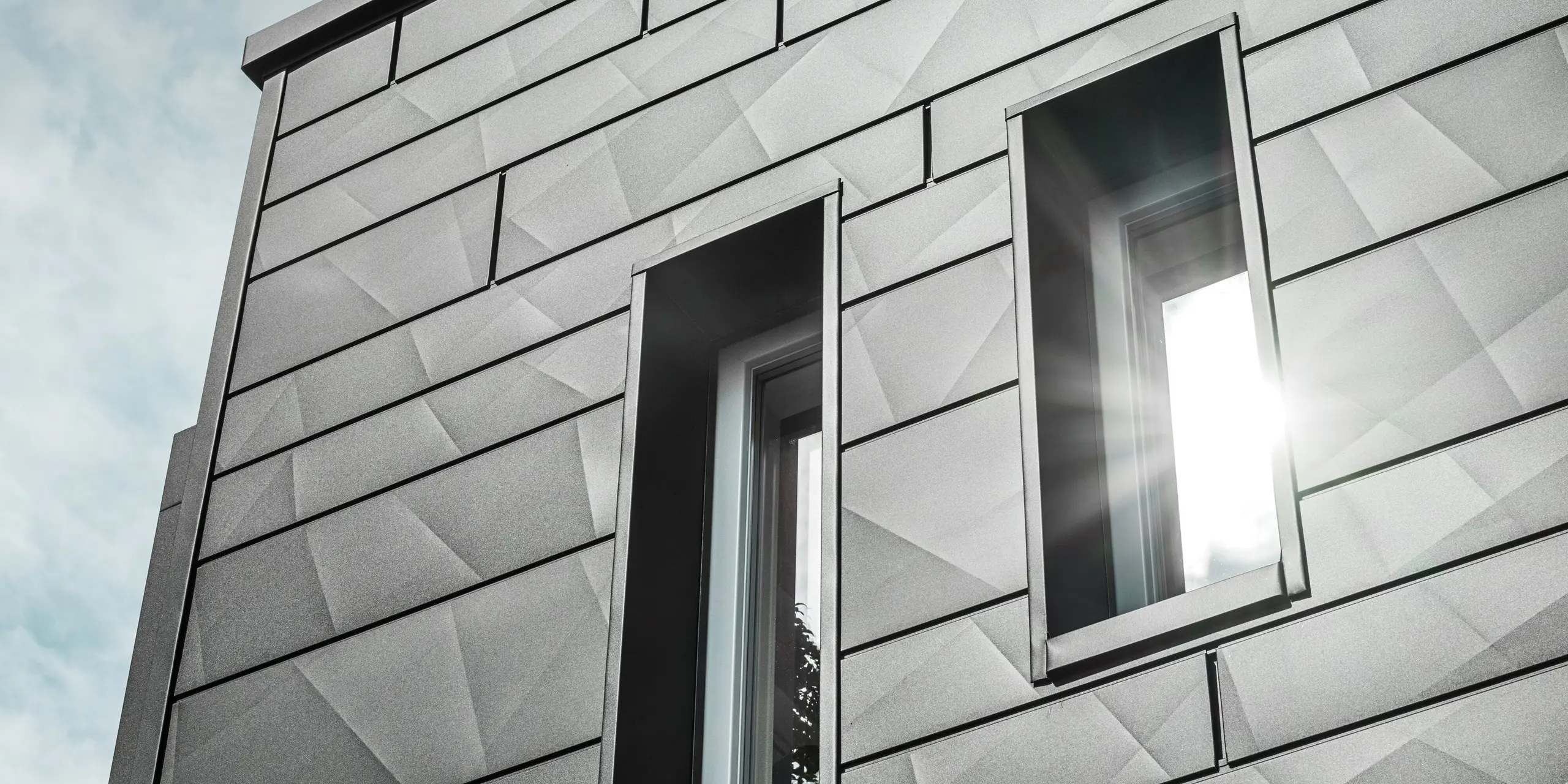 Moderná fasáda zrekonštruovaného rodinného domu v Teuschnitzi je pokrytá geometrickými fasádnymi panelmi PREFA Siding.X v čiernej farbe P.10. Okná s rámami z čiernych hliníkových kompozitných dosiek harmonicky zapadajú do celkového vzhľadu. Slnečný lúč preniká cez jedno z okien a zdôrazňuje čistý, štruktúrovaný povrch fasádnych panelov. Estetický dojem fasády umocňuje jemná hra svetla a materiálu.