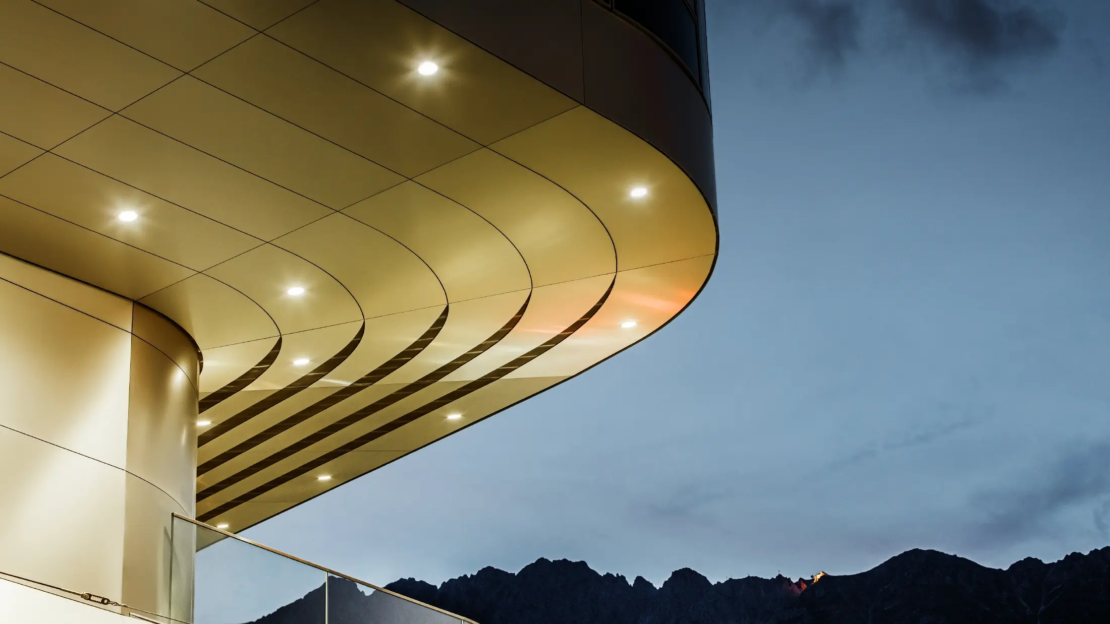 Obrázok ukazuje pôsobivý detailný pohľad na architektonický prvok z kompozitného panelu PREFA PEMA z lešteného hliníku. Panel je jemne zakrivený a má elegantné, plynulé línie, ktoré večer vyniknú vďaka cielenému osvetleniu. Čistá štruktúra a ušľachtilý vzhľad hliníka panelu dopĺňajú moderný dizajn fasády a ponúkajú elegantné, súčasné riešenie pre kvalitnú architektúru.