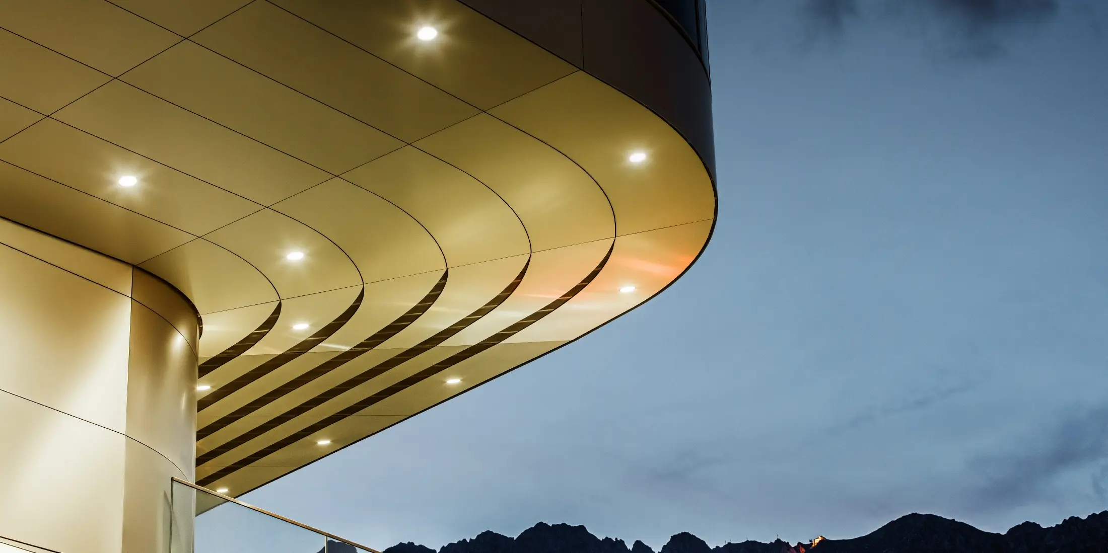Obrázok ukazuje pôsobivý detailný pohľad na architektonický prvok z kompozitného panelu PREFA PEMA z lešteného hliníku. Panel je jemne zakrivený a má elegantné, plynulé línie, ktoré večer vyniknú vďaka cielenému osvetleniu. Čistá štruktúra a ušľachtilý vzhľad hliníka panelu dopĺňajú moderný dizajn fasády a ponúkajú elegantné, súčasné riešenie pre kvalitnú architektúru.