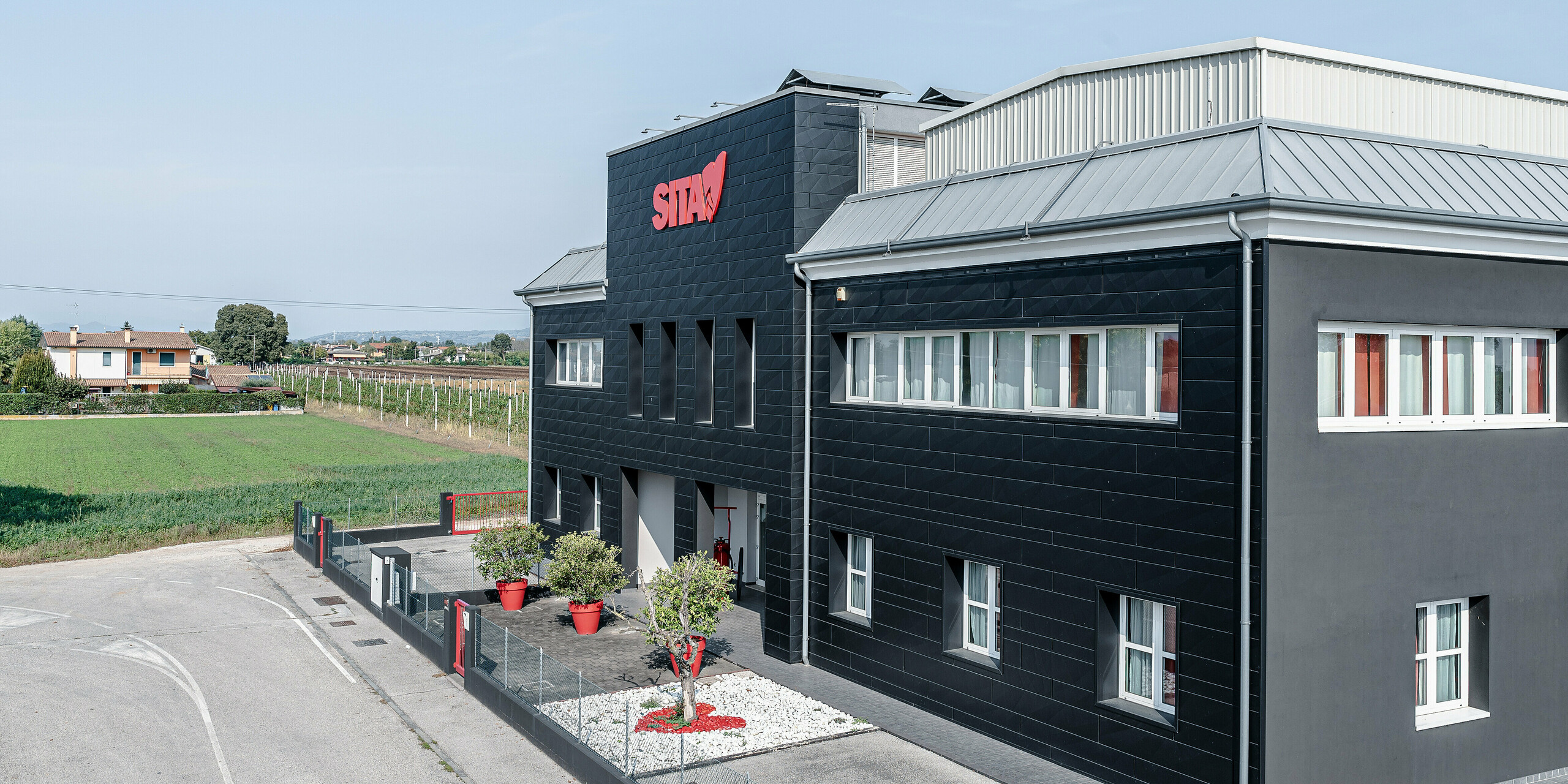 Budova spoločnosti SITA v talianskom Porcellengu uprostred idylickej krajiny, ktorú charakterizuje tmavá fasáda PREFA Siding.X vo farbe P.10 čierna. Fasáda je dokonalým príkladom úspešnej kombinácie modernej konštrukcie a zakomponovania do krajiny. Červené logo na budove nápadne vyniká na pozadí tmavých hliníkových panelov, ktoré sú nielen estetické, ale vďaka svojej odolnosti a udržateľnosti aj ideálne na priemyselné použitie. V pozadí vinič a polia dopĺňajú prírodnú atmosféru a zdôrazňujú miestne korene spoločnosti.