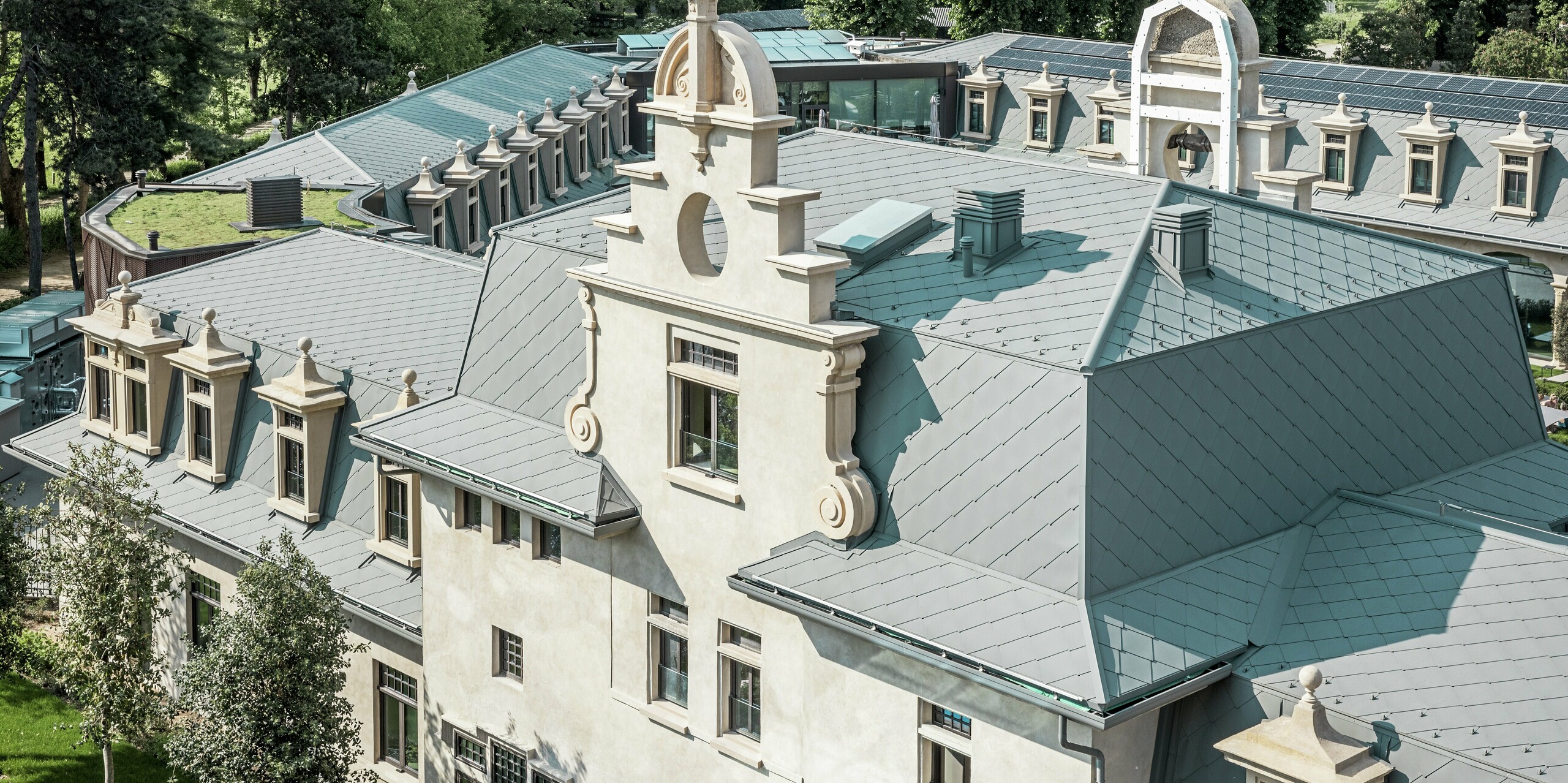 Detailný pohľad na strechu termálneho parku „De Montel – Terme Milano“ s precízne vykonanou krytinou z PREFA strešných šablón 44 × 44 v odtieni P.10 svetlošedá. Zložitá geometria strechy s množstvom vikierov, ríms, štítov a ventilačných prvkov je majstrovsky vyriešená. Historickú architektúru harmonicky dopĺňajú moderné prvky, ako je zelená plochá strecha a bezšvíkovo integrovaná fotovoltická inštalácia v pozadí.