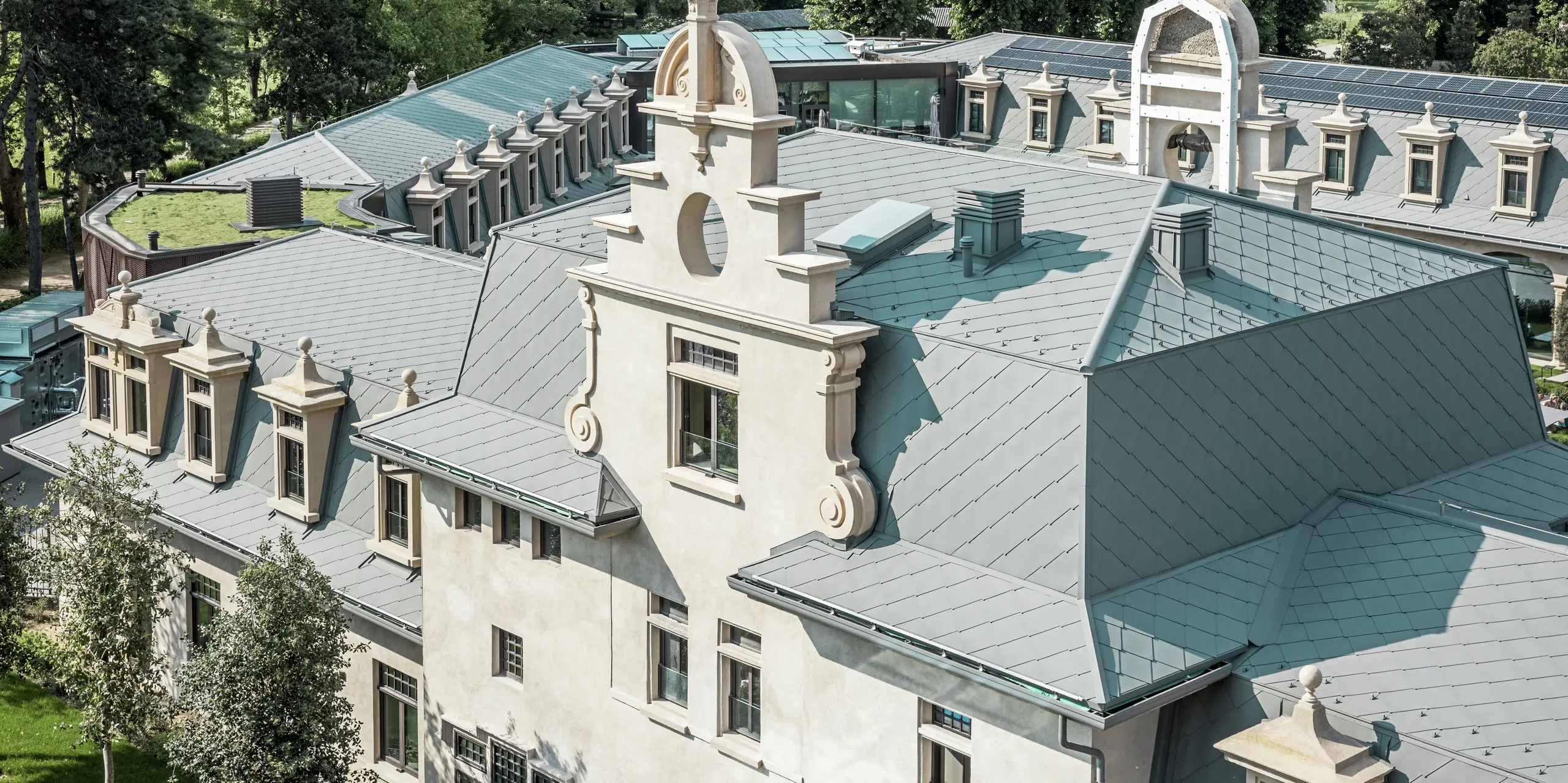 Detailný pohľad na strechu termálneho parku „De Montel – Terme Milano“ s precízne vykonanou krytinou z PREFA strešných šablón 44 × 44 v odtieni P.10 svetlošedá. Zložitá geometria strechy s množstvom vikierov, ríms, štítov a ventilačných prvkov je majstrovsky vyriešená. Historickú architektúru harmonicky dopĺňajú moderné prvky, ako je zelená plochá strecha a bezšvíkovo integrovaná fotovoltická inštalácia v pozadí.