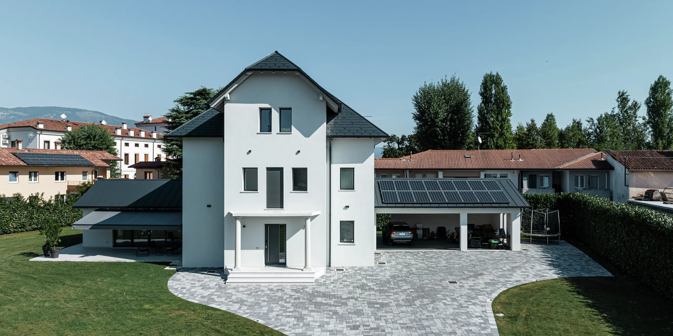 Pohľad z prednej strany na moderný rodinný dom v Marano Vicentino v Taliansku s odolnými strešnými šindľami PREFA P.10 v antracitovej farbe. Susedné vedľajšie budovy vľavo a vpravo sú pokryté osvedčeným dvojitým stojatým falcom z hliníka – tiež v antracitovej farbe. Nad garážou zabezpečuje fotovoltická inštalácia elektrickú energiu z obnoviteľných zdrojov. Svetlá omietková fasáda a tmavá strecha vytvárajú elegantný kontrast na trojposchodovom rodinnom dome.