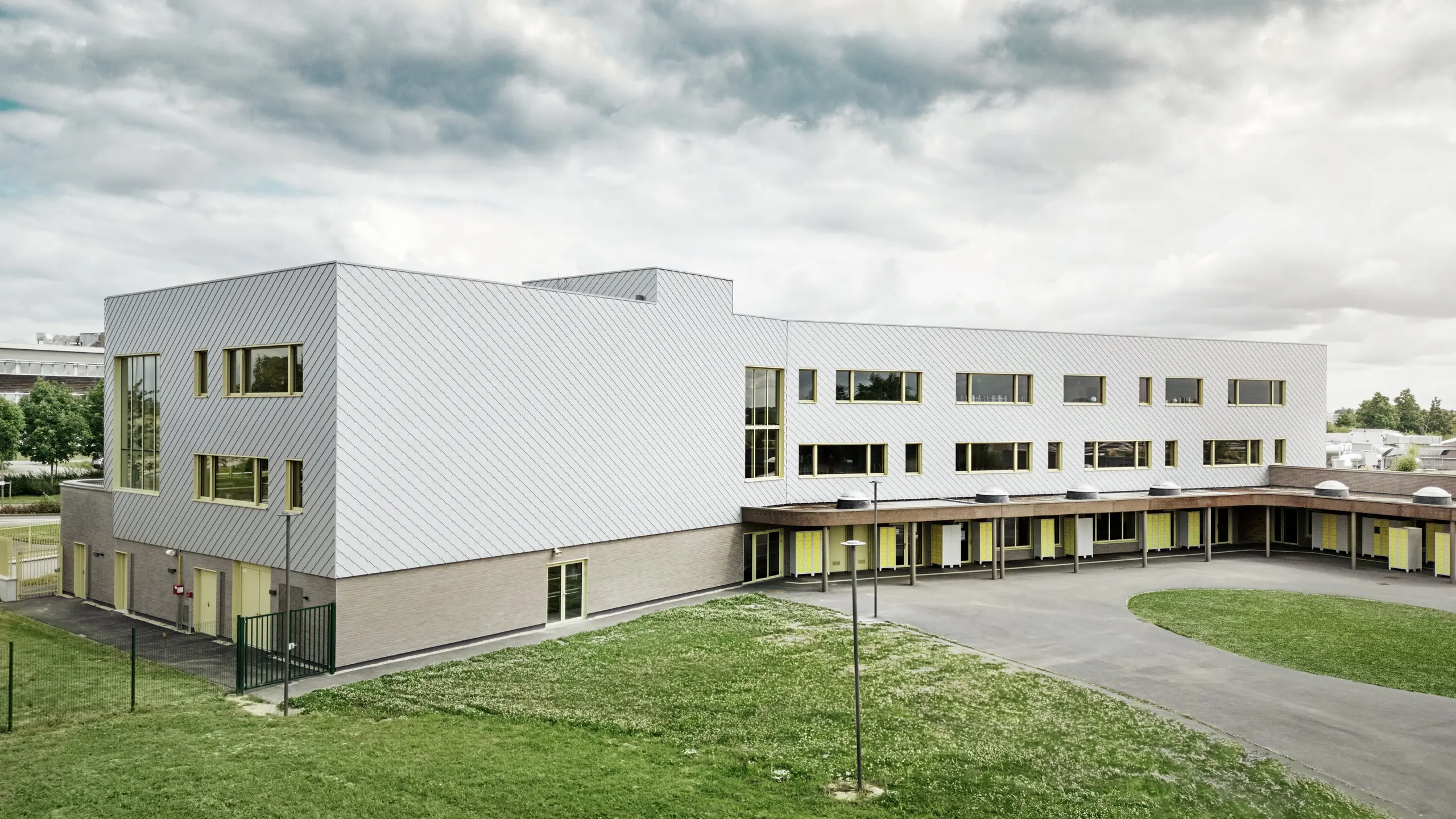 Na obrázku je zadná časť školy Collège Léontine Dolivet. V popredí sa nachádza veľké asfaltové školské ihrisko s jasnými líniami a otvorenými plochami, ktoré sa dajú využiť na prestávky a voľnočasové aktivity. Niekoľko zelených plôch oživuje okolie. V pozadí sa týči moderná budova školy s výraznou hliníkovou fasádou z fasádnych šablón, ktoré odrážajú svetlo a vytvárajú dynamický efekt. Niekoľko veľkých okien zaisťuje svetlosť v interiéri. Scéna je nasnímaná za denného svetla, pri mierne zamračenom nebi, čo zvlášť zdôrazňuje kovový povrch.