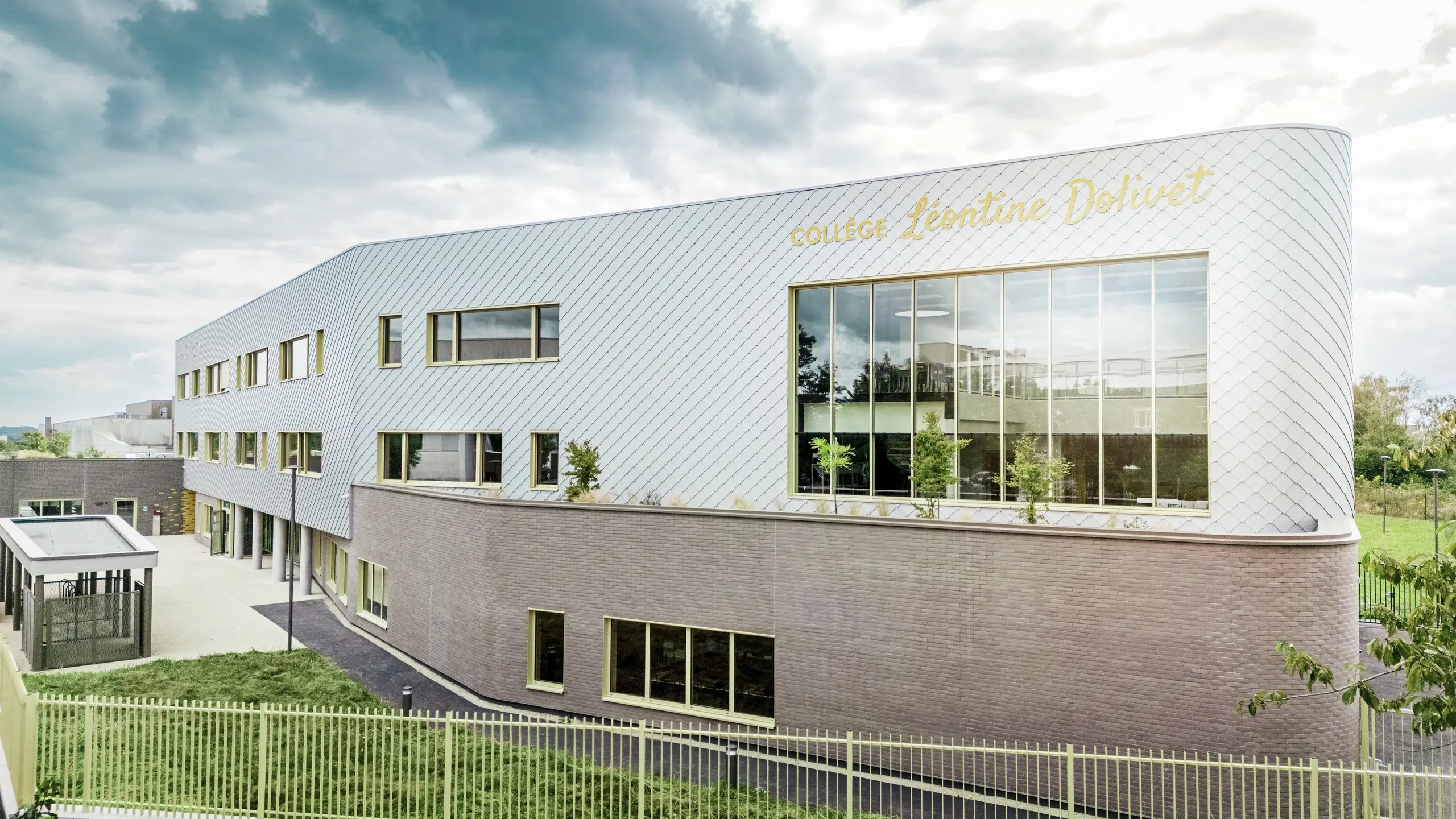 Bočný pohľad na Collège Léontine Dolivet v Cesson-Sévigné s modernou hliníkovou fasádou z PREFA fasádnych šablón v striebornej metalickej farbe. Diagonálna štruktúra vytvára dynamický vzhľad, zatiaľ čo veľké sklenené plochy a žlté okenice dodávajú budove zaujímavé akcenty. V popredí sú viditeľné zelené plochy a plot, ktoré rámujú budovu. Trvanlivé fasádne riešenie PREFA spája eleganciu, odolnosť voči poveternostným vplyvom a udržateľnosť – architektonický pohľad upútavajúci jasnými líniami a meniacim sa kovom.
