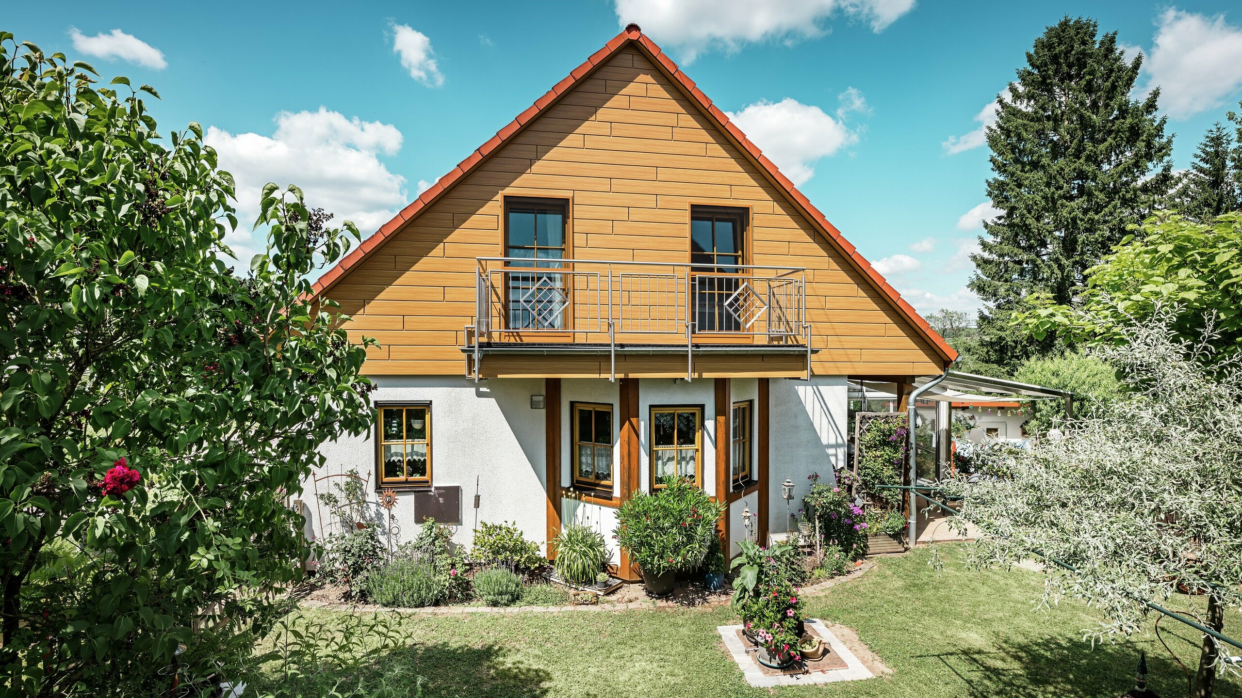 Na tejto fotografii žiari zrekonštruovaná štítová stena rodinného domu v Blieskasteli v teplom odtieni dubu prírodného vďaka Sidingom PREFA. Vysoko kvalitná hliníková fasáda s realistickým vzhľadom dreva vytvára výrazný vizuálny akcent a pôsobivo ladí s červenou škridlovou strechou a bujnou záhradou. Obklopený kvetinami, kríkmi a sviežou zeleňou pôsobí dom prívetivo a udržiavane. Povrch PREFA kombinuje prirodzený vzhľad s funkčnými výhodami, ako je vysoká odolnosť voči poveternostným vplyvom, stabilita a takmer bezúdržbová životnosť – estetický vrchol s trvalou pridanou hodnotou.