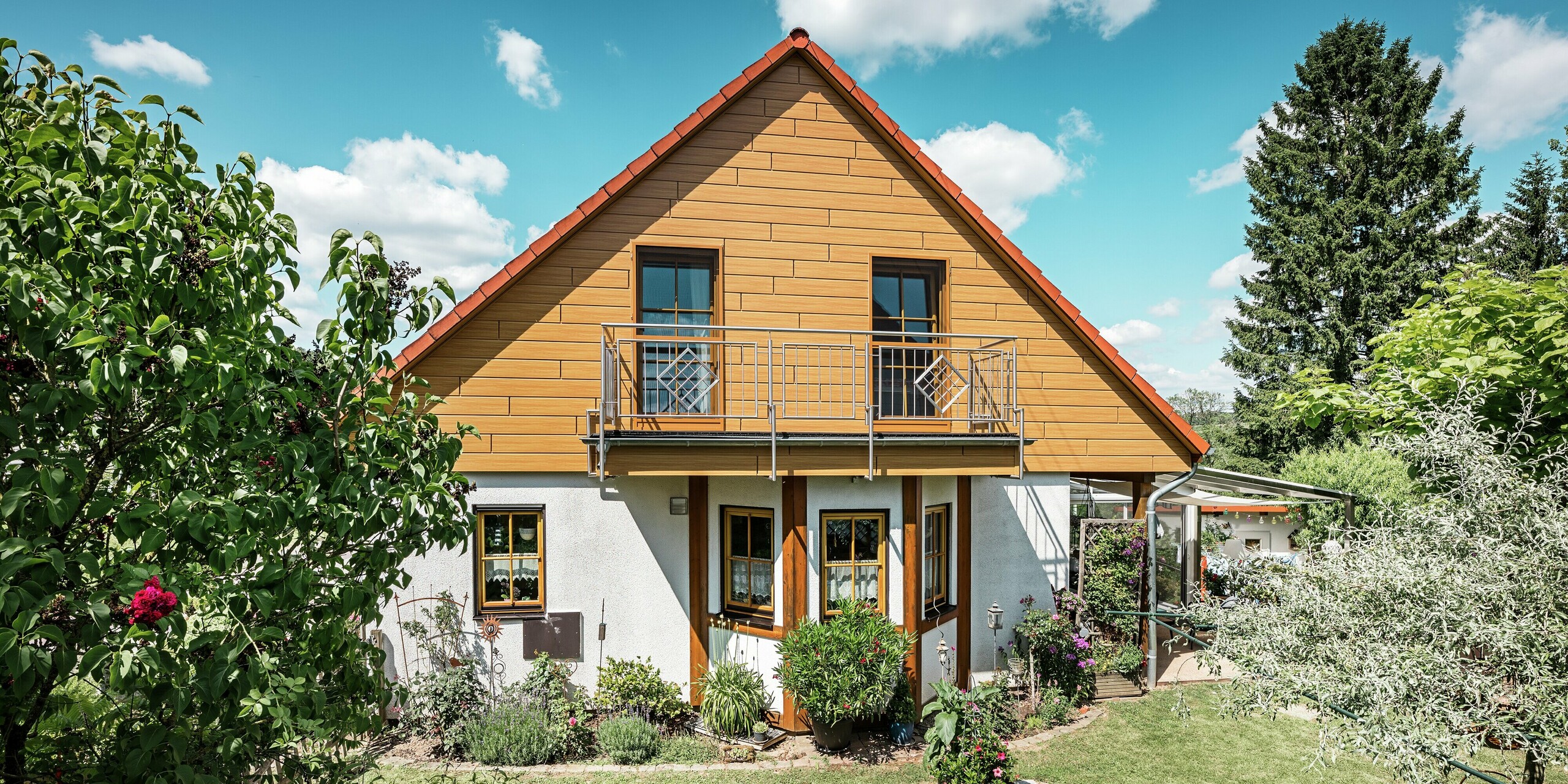 Auf diesem Bild strahlt die sanierte Giebelseite eines Einfamilienhauses in Blieskastel in warmem Eiche-Natur-Ton dank der PREFA Sidings. Die hochwertige Aluminiumfassade in realistischer Holzoptik setzt einen starken visuellen Akzent und harmoniert eindrucksvoll mit dem roten Ziegeldach und dem üppigen Garten. Umgeben von Blumen, Sträuchern und sattem Grün wirkt das Haus einladend und gepflegt. Die PREFA Oberfläche kombiniert natürliche Optik mit funktionalen Vorteilen wie hoher Witterungsbeständigkeit, Stabilität und nahezu wartungsfreier Langlebigkeit – ein ästhetisches Highlight mit nachhaltigem Mehrwert.