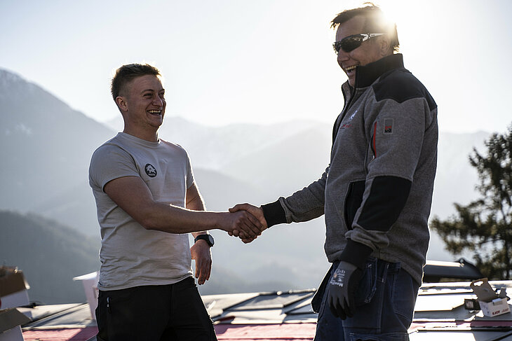 Na šikmej streche domu stojí školiteľ spoločnosti PREFA a klampiar, ktorí si podávajú ruky. Obaja majú na sebe profesionálne pracovné oblečenie a bezpečnostnú výstroj. V pozadí je vidieť nízko stojace slnko a obaly hliníkových výrobkov PREFA. Táto scéna symbolizuje úspešnú spoluprácu a odbornú výmenu s kvalitou potvrdenou potrasením ruky. Školiteľ spoločnosti PREFA podporuje pokrývača svojimi znalosťami v oblasti spracovania, techniky a produktových systémov. Obrázok symbolizuje úzku podporu remeselníkov zo strany spoločnosti PREFA – od školenia až po profesionálnu realizáciu na stavbe.