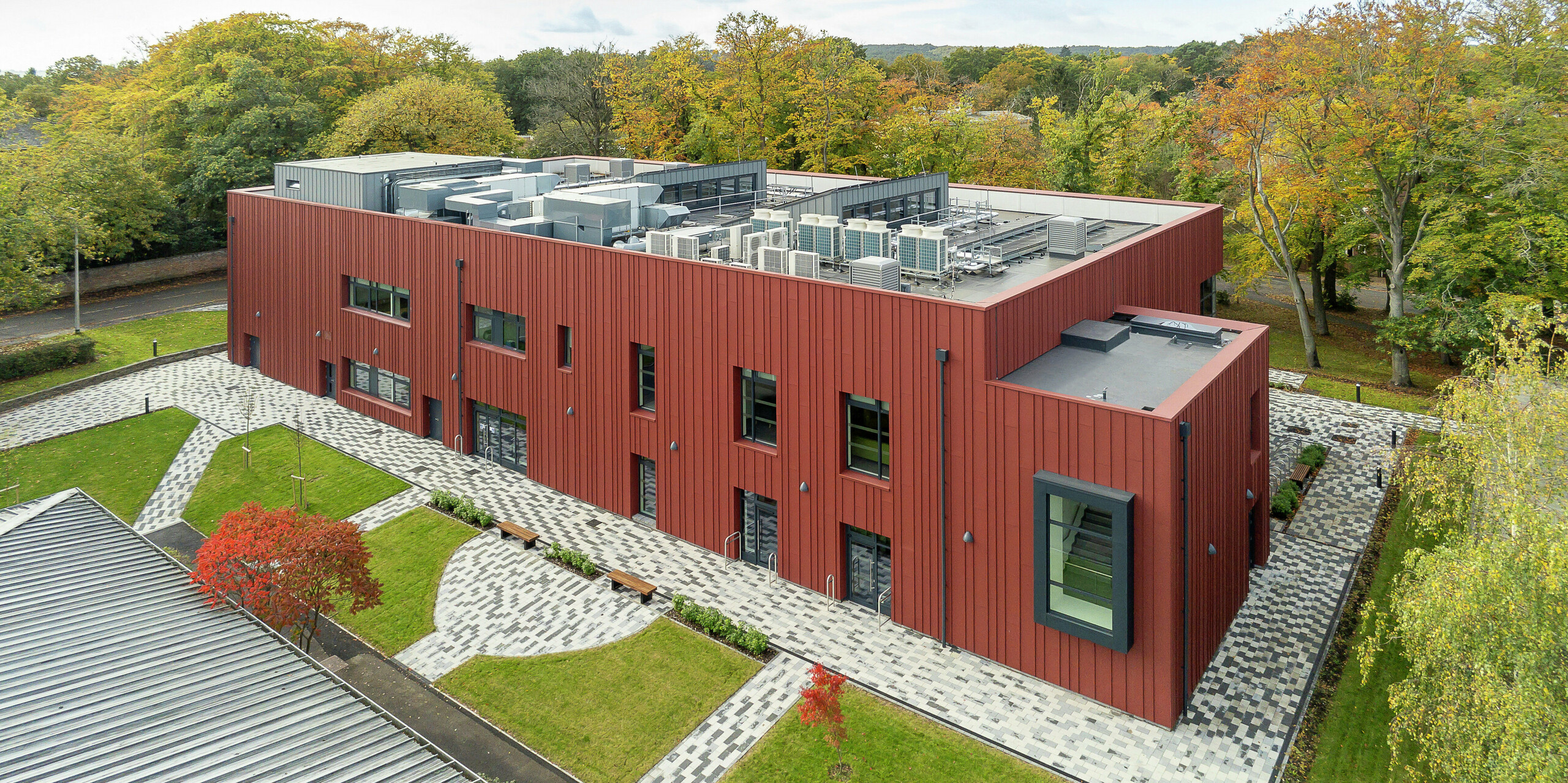 Architektúra v súlade s prírodou: Soldiers' Centre v Pirbright z vtáčej perspektívy. Výrazná stojatá fasáda z PREFALZu v odtieni P.10 tmavočervená sa harmonicky začleňuje do zeleného prostredia. Vystupujúce okenné rámy v P.10 Anthrazit dodávajú moderný akcent. Štruktúrované vonkajšie priestory s dláždenými chodníkmi, zelenými plochami a miestami na sedenie vytvárajú príjemnú atmosféru. Na plochú strechu sú integrované technické inštalácie, ktoré prispievajú k modernej technike budovy.