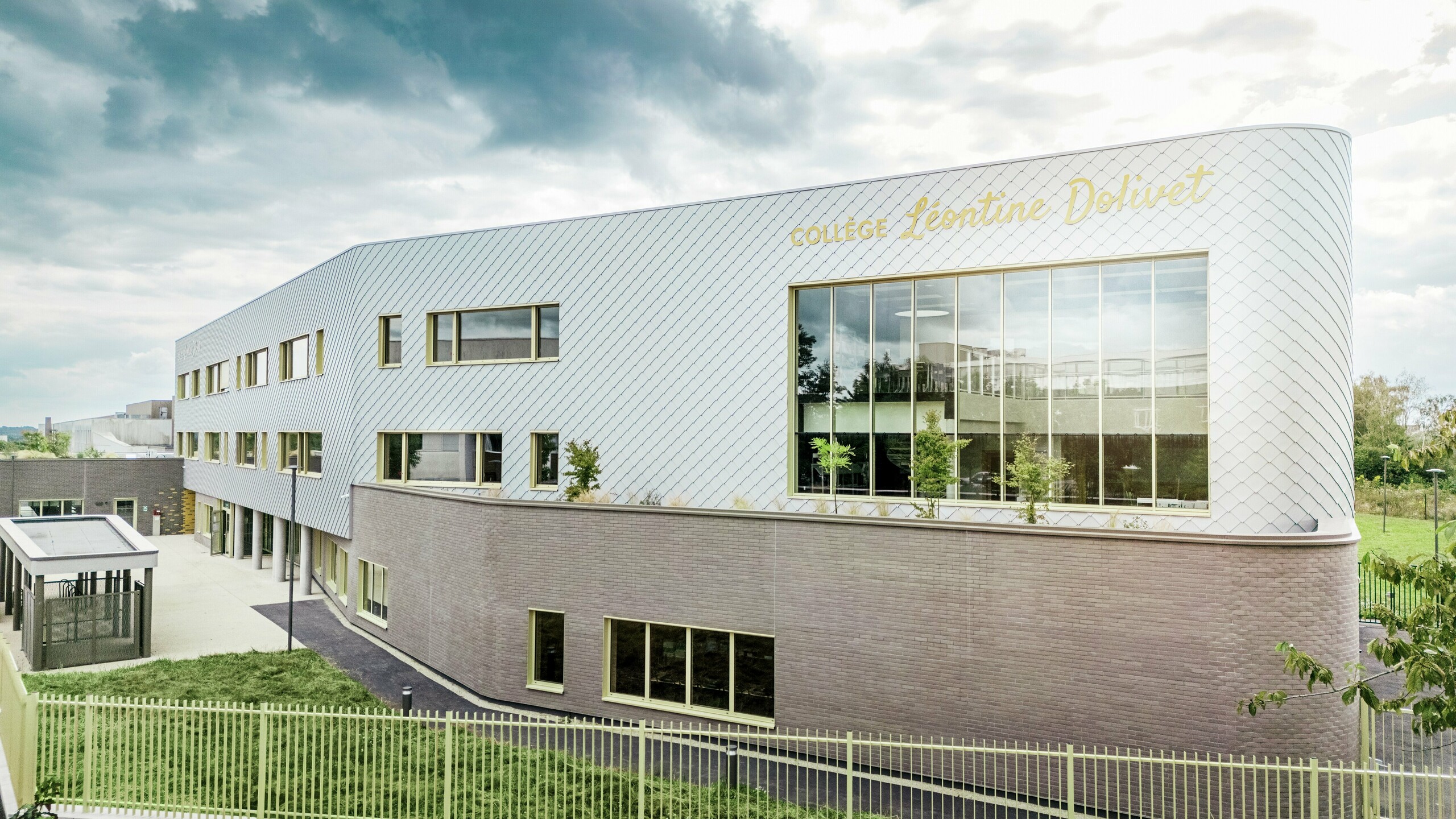 Bočný pohľad na Collège Léontine Dolivet v Cesson-Sévigné s modernou hliníkovou fasádou z PREFA fasádnych šablón v striebornej metalickej farbe. Diagonálna štruktúra vytvára dynamický vzhľad, zatiaľ čo veľké sklenené plochy a žlté okenice dodávajú budove zaujímavé akcenty. V popredí sú viditeľné zelené plochy a plot, ktoré rámujú budovu. Trvanlivé fasádne riešenie PREFA spája eleganciu, odolnosť voči poveternostným vplyvom a udržateľnosť – architektonický pohľad upútavajúci jasnými líniami a meniacim sa kovom.