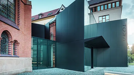Obrázok ukazuje modernú prístavbu knižnice, ktorá elegantne nadväzuje na historický Červený kostol v Olomouci. Výrazná novostavba zaujme fasádou z čiernych kompozitných dosiek PREFABOND z hliníka, ktorá je navrhnutá v jasných geometrických tvaroch a presvedčí precíznou prácou s detailmi. Sklenené steny budovy ponúkajú transparentný pohľad do interiéru, ktorý harmonicky zapadá do historického prostredia. Súhra moderných materiálov a klasických architektonických prvkov vytvára zaujímavé vizuálne prepojenie medzi starým a novým. Inovatívne použitie dosiek PREFABOND zaisťuje nielen moderný vzhľad, ale aj dlhú životnosť a odolnosť, ktoré budovu pripravujú na budúcnosť.
