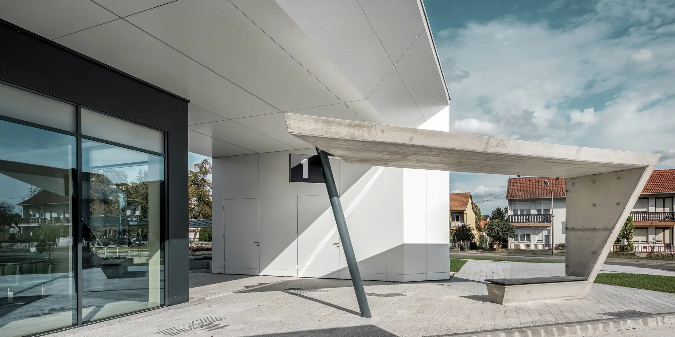 Vstupná časť autobusovej stanice Lenti; spodná strana strechy je obložená hliníkovým kompozitným panelom PREFABOND od spoločnosti PREFA v čiste bielej farbe, fasádne prvky pri vchodových dverách a výkladoch sú obložené v antracitovej farbe P.10.