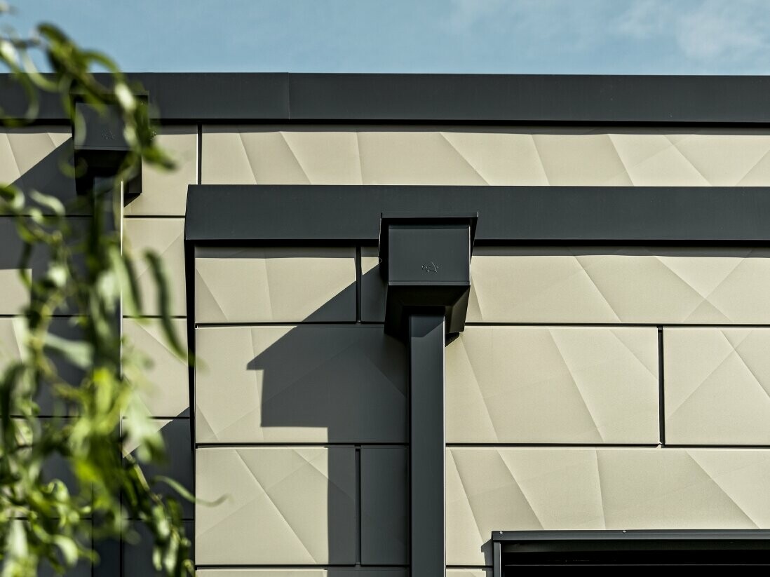 Moderný rodinný dom s plochou strechou a úzkymi oknami. Fasáda je obložená panelmi PREFA Siding.X, fasáda s nariaseným vzhľadom v bronzovej farbe. Napojenie bolo zhotovené v antracitovej farbe. Na odvodnenie strechy slúži hranatý zvod PREFA s vhodnými zbernými kotlíkmi v antracitovej farbe.