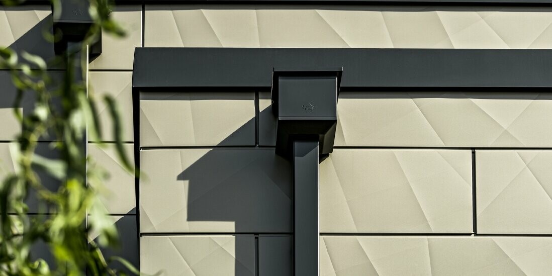 Moderný rodinný dom s plochou strechou a úzkymi oknami. Fasáda je obložená panelmi PREFA Siding.X, fasáda s nariaseným vzhľadom v bronzovej farbe. Napojenie bolo zhotovené v antracitovej farbe. Na odvodnenie strechy slúži hranatý zvod PREFA s vhodnými zbernými kotlíkmi v antracitovej farbe.