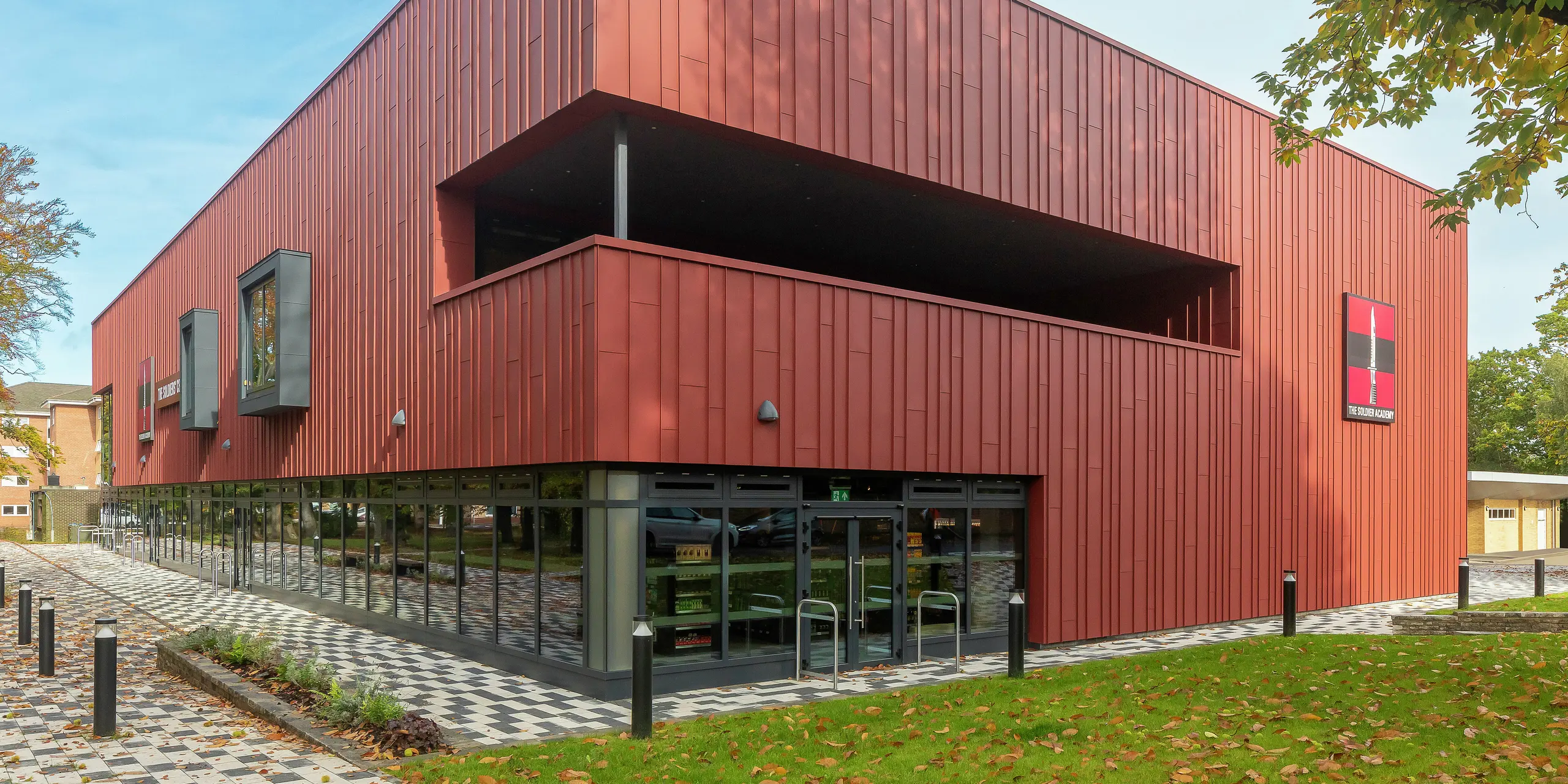 Moderná architektúra s PREFALZ: Soldiers' Centre v Pirbrightu zaujme modernou stojatou falcovou fasádou z PREFALZ v odtieni P.10 tmavočervená. Rozsiahle zasklenie na prízemí vytvára transparentnosť a otvorenosť, zatiaľ čo okenné rámy v P.10 Anthrazit dodávajú štýlové akcenty. Udržateľný stavebný koncept s odolnými hliníkovými produktmi PREFA pre najvyššie nároky na dizajn a kvalitu.