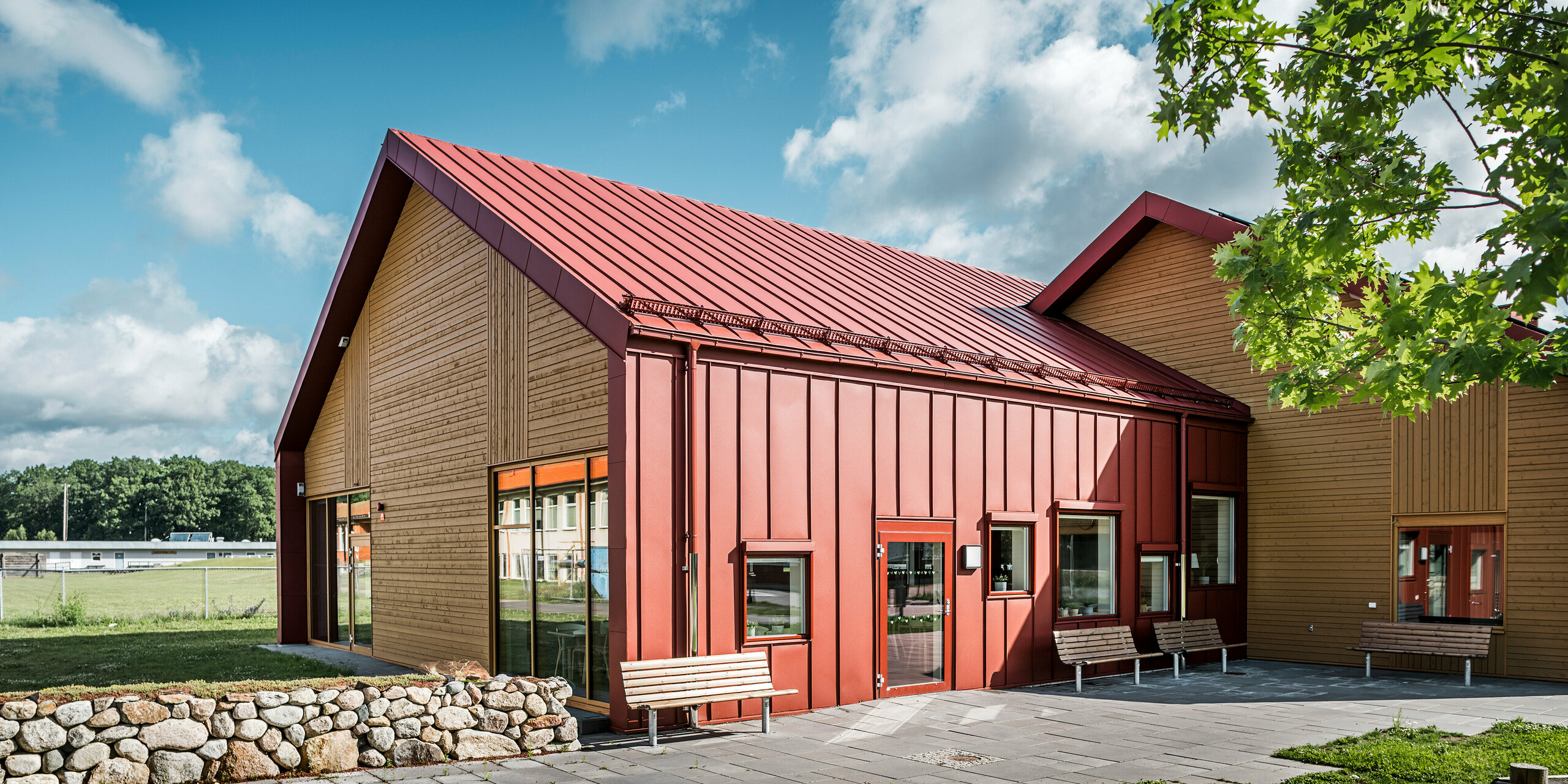 Pohľad na exteriér školskej jedálne Restaurang Gröna Hjärtat v Broby, Švédsko. Na snímke sú lavičky pred budovou, ktorá má na štítovej strane nápadnú kombináciu tmavočervenej farby PREFALZ a dreva. Architektonický návrh zdôrazňuje jasné línie a harmonický kontrast medzi červenými hliníkovými povrchmi a teplým tónom dreva. V popredí možno vidieť kamenné múry a zelené plochy, ktoré rámcujú moderný dizajn budovy. Okolie a budova sa spájajú do príjemného a premysleného priestoru.