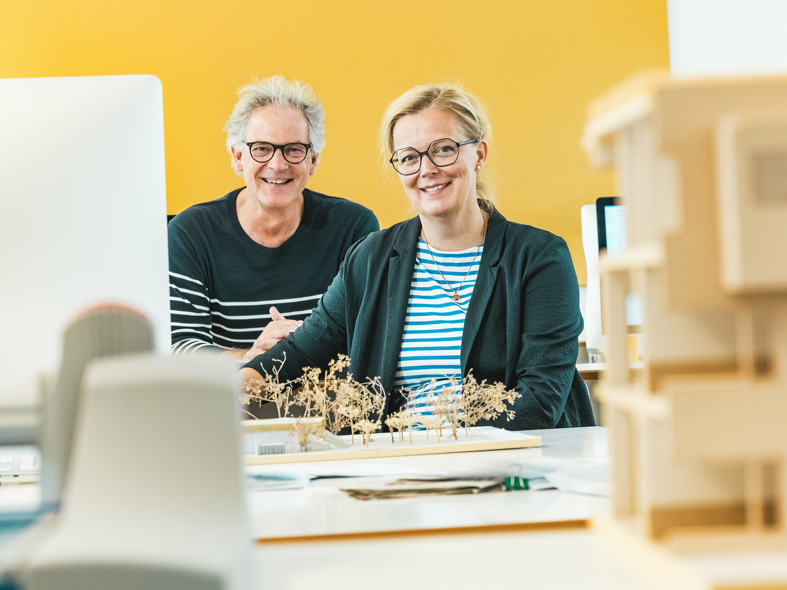 Architektka Claudia a architekt Theo Härtner s úsmevom sedia pred architektonickým modelom. Tento architektonický pár realizuje materskú školu v Alzenau, perspektívnu hybridnú budovu s fasádou z hliníka PREFA. V popredí modelu sú miniatúrne stromčeky, ktoré odrážajú udržateľný celkový koncept. Materská škola v Alzenau spája inovatívnu architektúru s zodpovedným mestským rozvojom a ukazuje všestrannosť bronzových PREFA fasádnych šablón 44 × 44. Trvácna, nenáročná na údržbu a plne recyklovateľná hliníková fasáda vizuálne prepája obytnú časť a časť pre materskú školu do harmonickej jednoty. Obrázok zachytáva kreatívnu energiu plánovacieho procesu a zdôrazňuje estetické a ekologické výhody produktov PREFA.