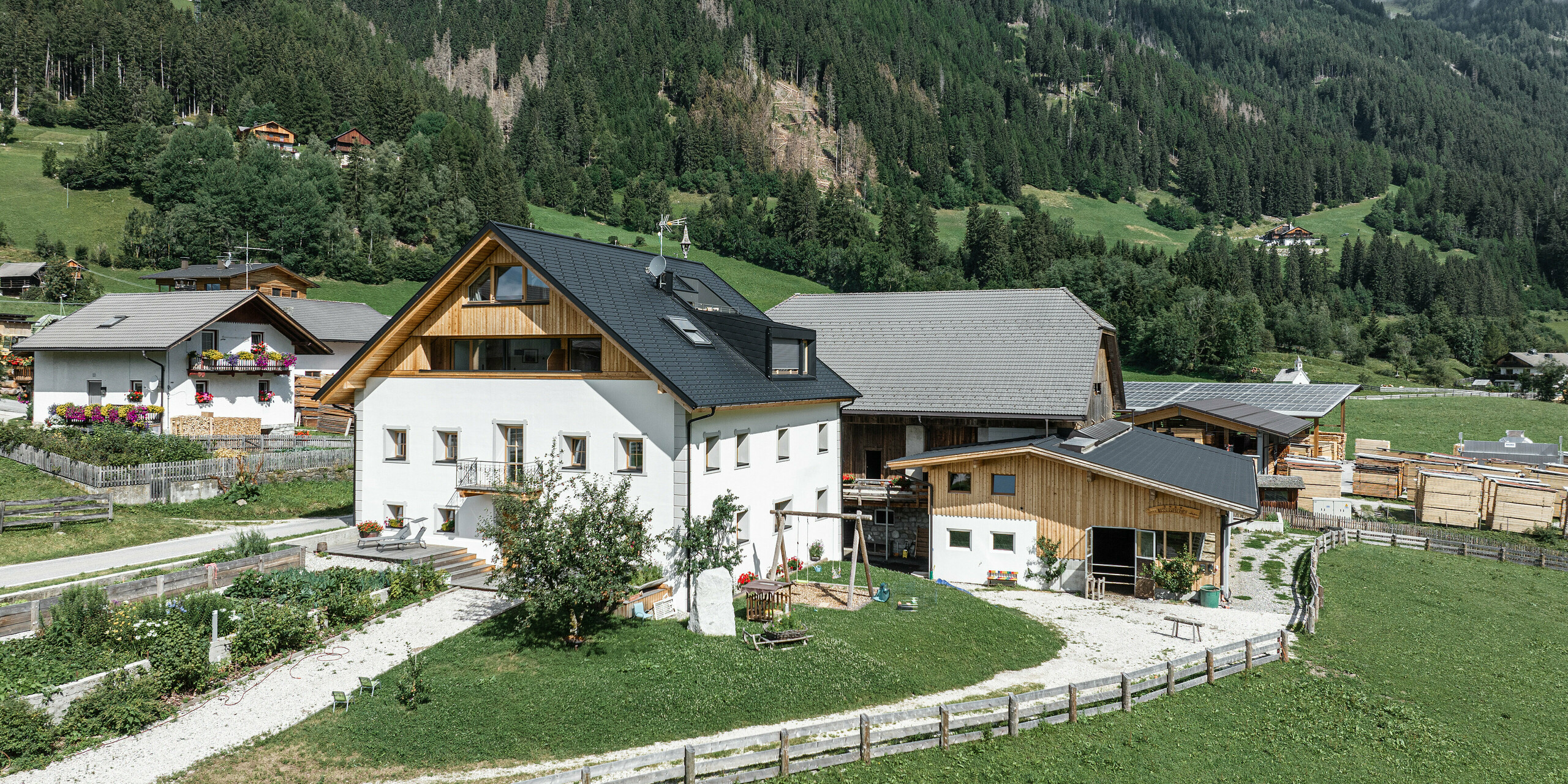 Pohľad z vtáčej perspektívy na Ausserwegerhof v Južnom Tirolsku so strechou PREFA vo farbe P.10 antracitová. Dovolenkové apartmány sú zasadené do zelenej krajiny údolia Antholz s malebnými lesmi a horami v pozadí. Robustný strešný šindeľ PREFA DS.19 chráni tradičné stavby ako v teplých letných mesiacoch, tak aj v ľadových zimných podmienkach.