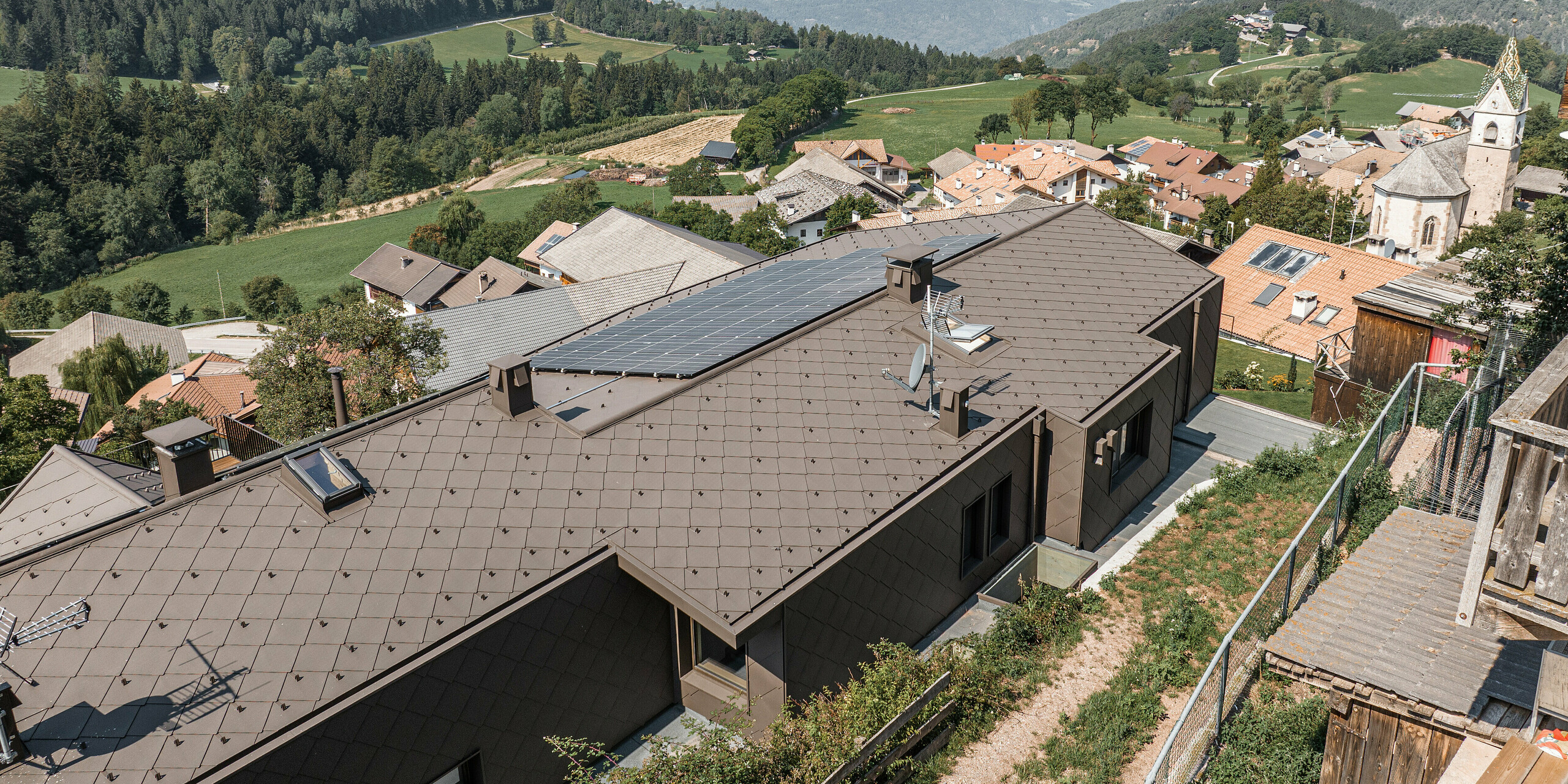 Letecký pohľad na modernizovaný dvojdom v juhotirolskom Möltene so strechou z hnedých hliníkových šablón PREFA a fotovoltického systému. Dom bez problémov zapadá do prostredia dediny s tradičnými budovami a kostolom na pozadí širokého údolia a zelených hôr. Odolné hliníkové šablóny poskytujú ako funkčnú ochranu, tak príťažlivú estetiku v súlade s prírodou.